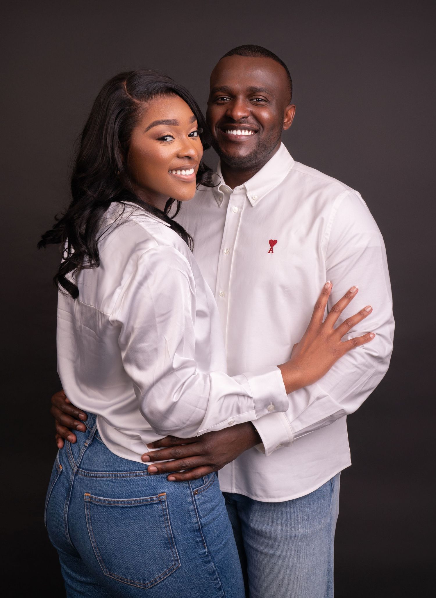 A couple in matching white button-down shirts and jeans embrace and smile warmly against a dark backdrop.