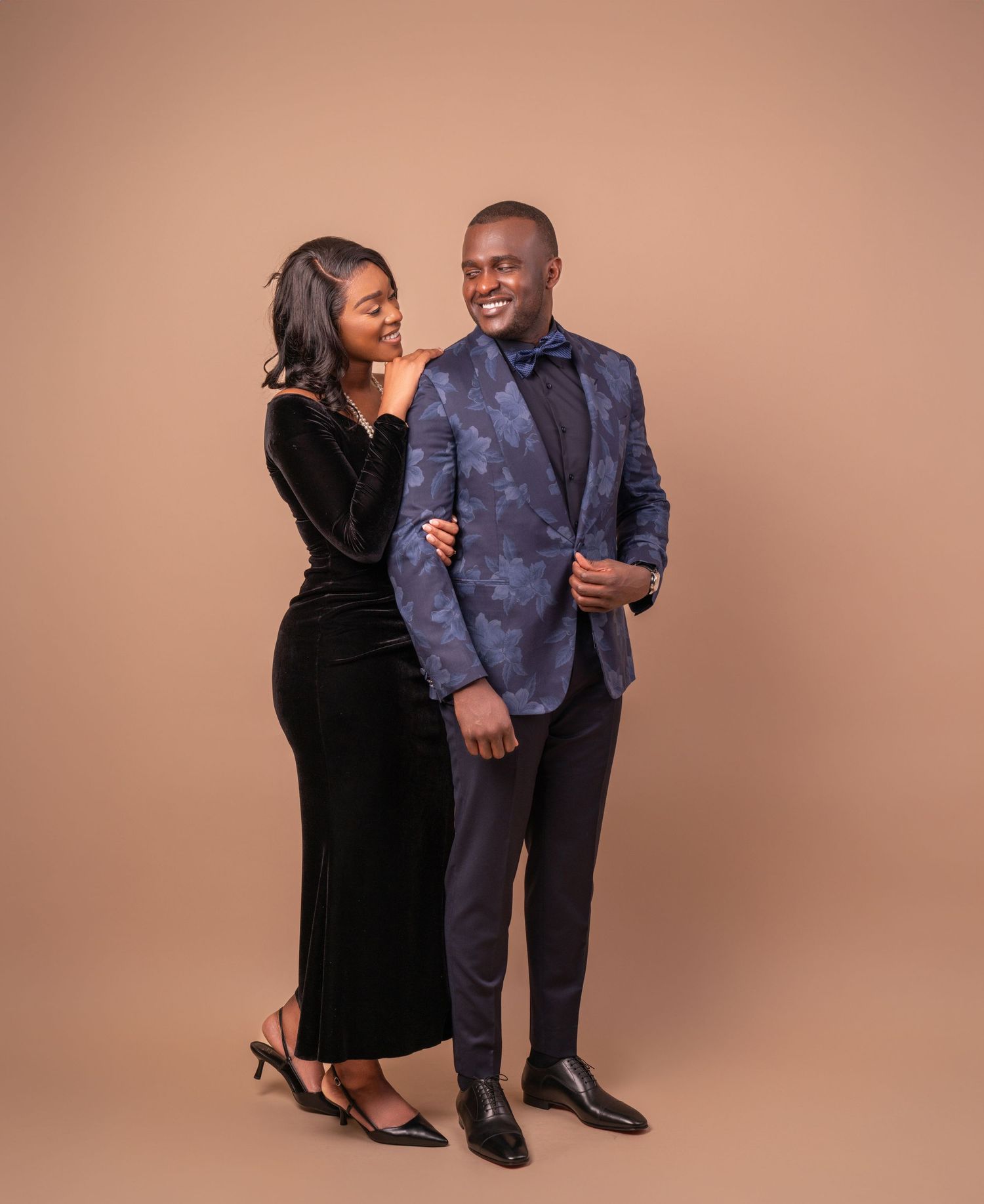 A couple poses together against a brown backdrop wearing formal attire - a black velvet dress and gray patterned blazer.