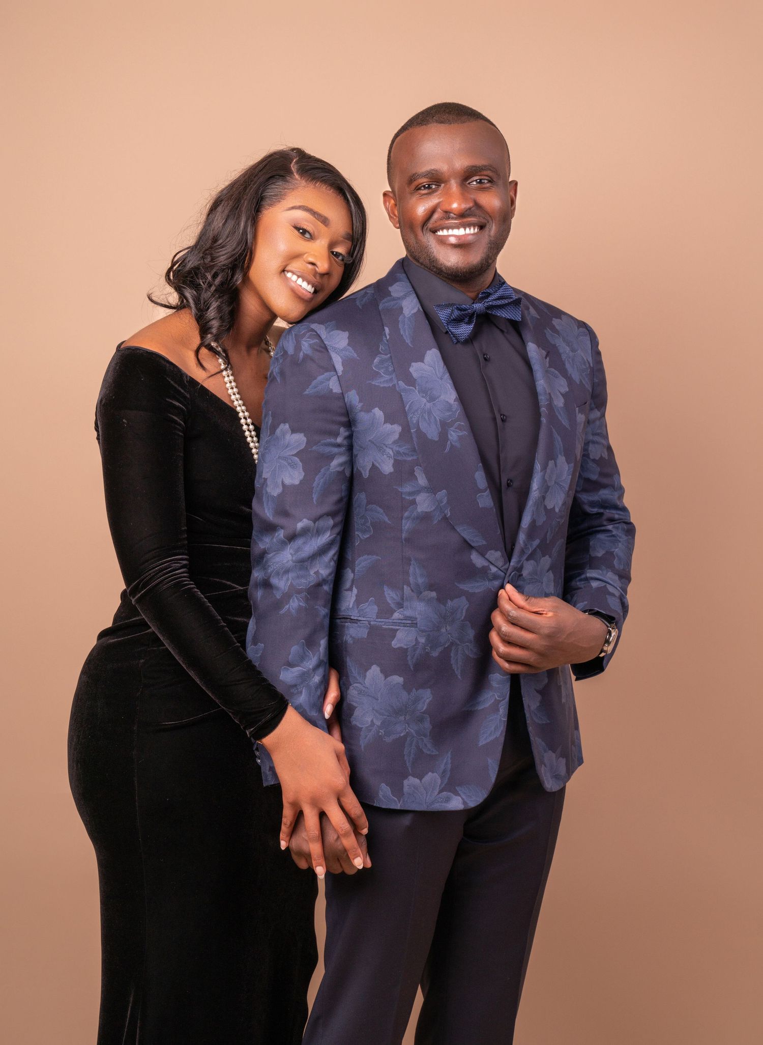 A couple poses in formal attire, wearing a floral blue blazer and black velvet dress against a pink background.