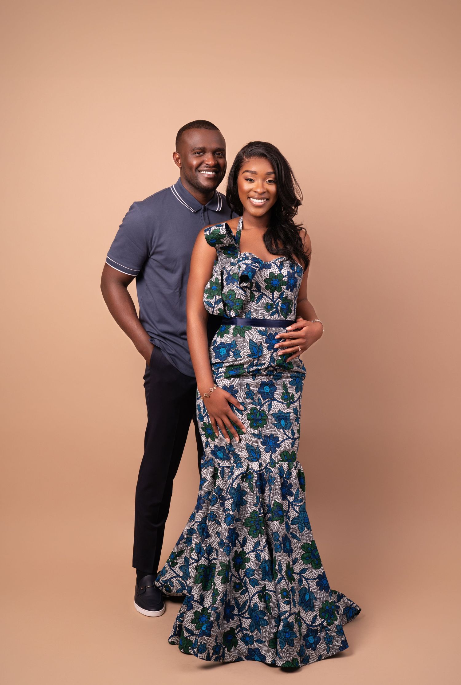 A couple poses together against a beige backdrop, with one wearing a blue polo and the other in a floral maxi dress.