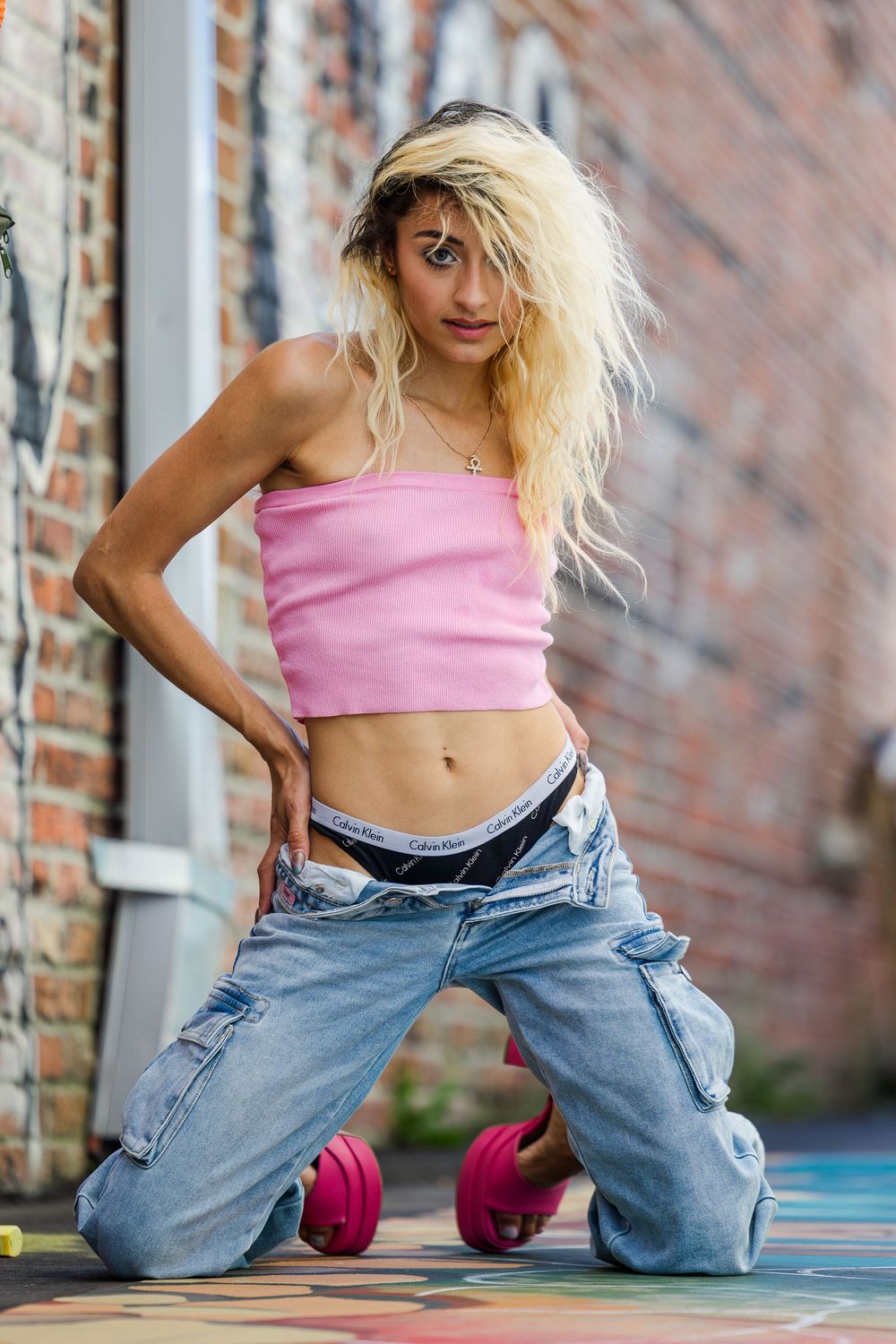Blonde woman in pink crop top and low-rise cargo jeans posing against brick wall.