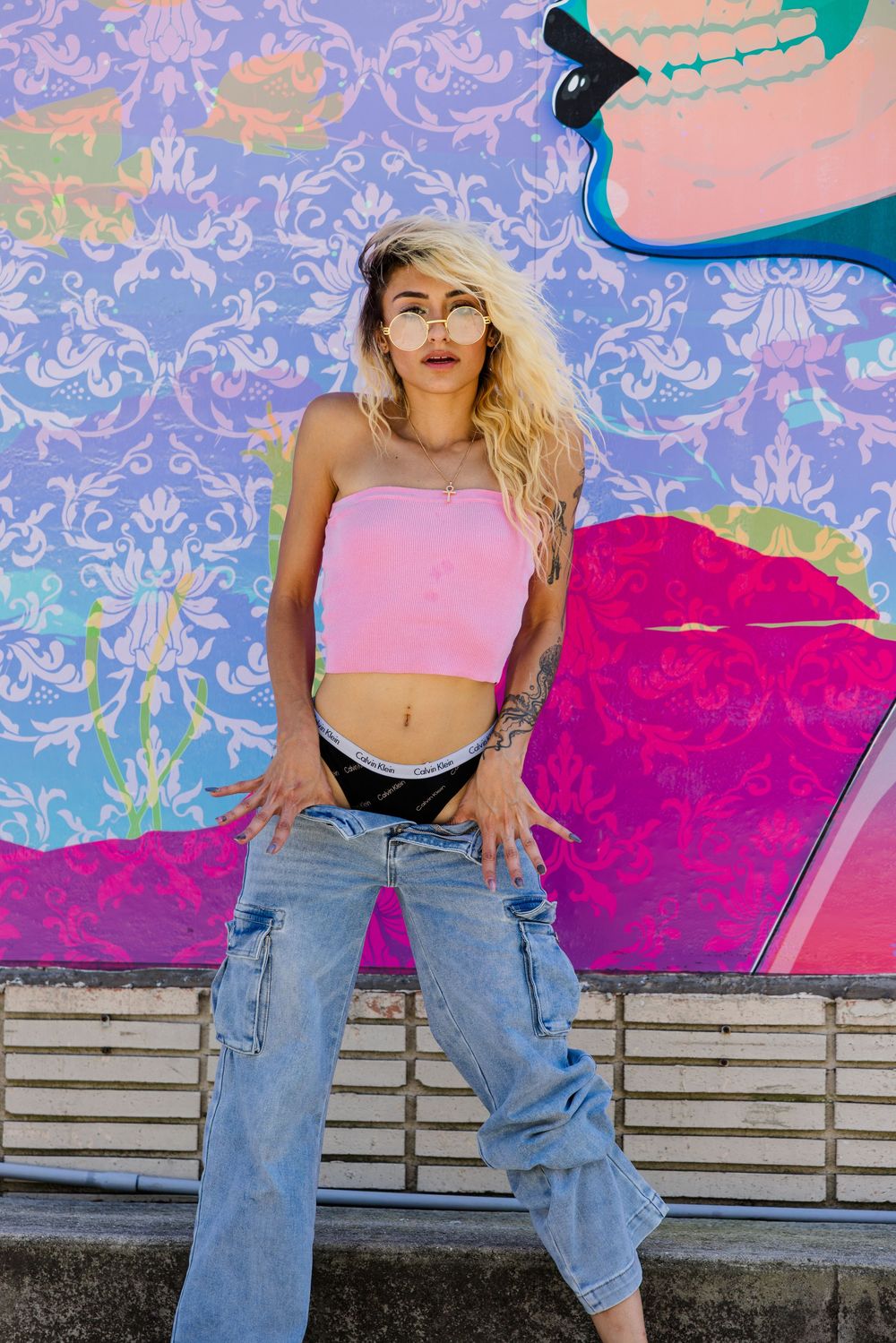 Blonde woman in pink tube top and baggy jeans posing against vibrant graffiti mural.