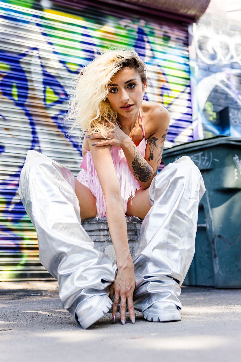 Blonde tattooed woman in pink top and silver pants sitting near graffiti-covered garage.