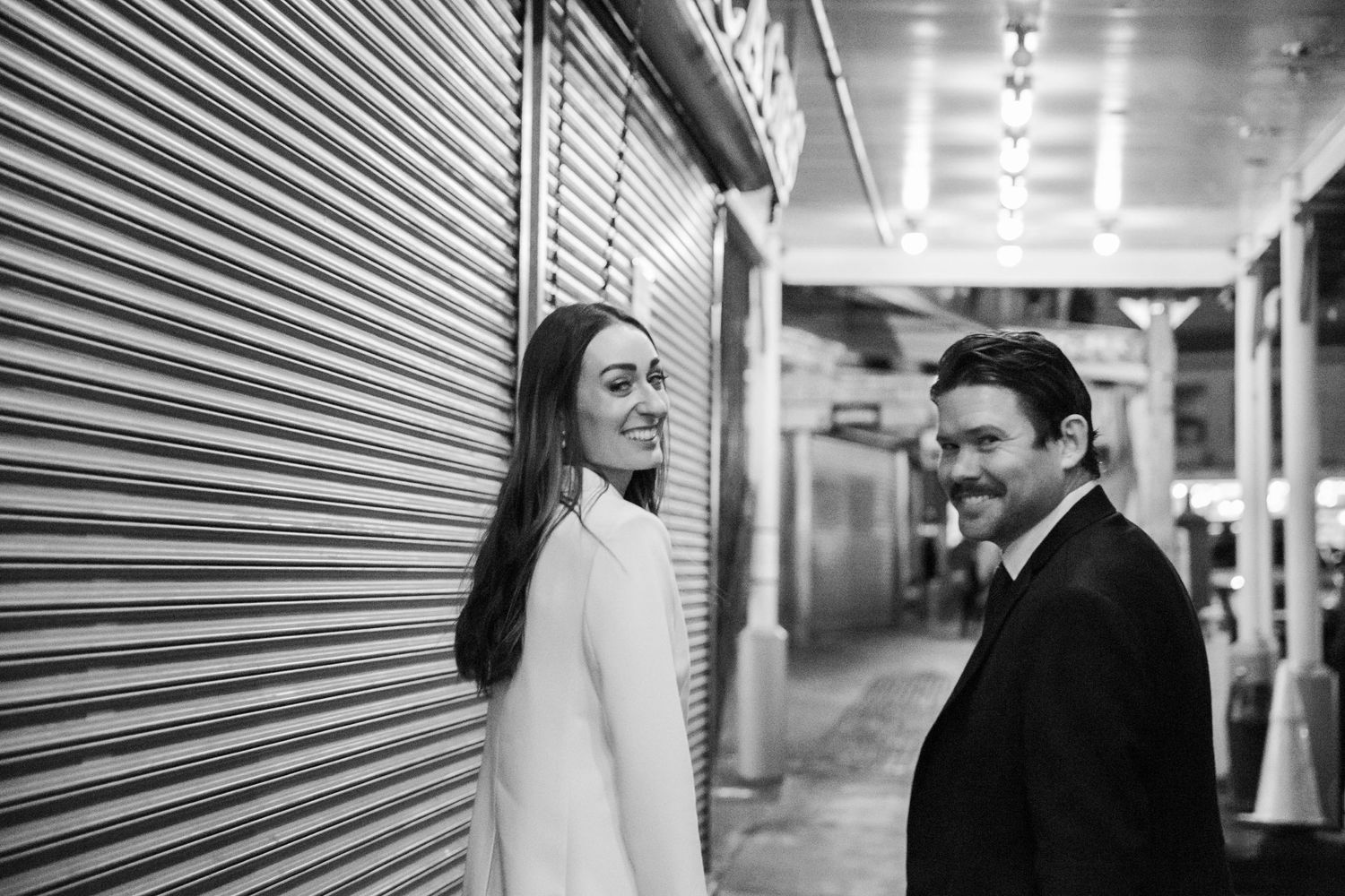 A couple shares a playful moment in an urban corridor with industrial metal shutters.