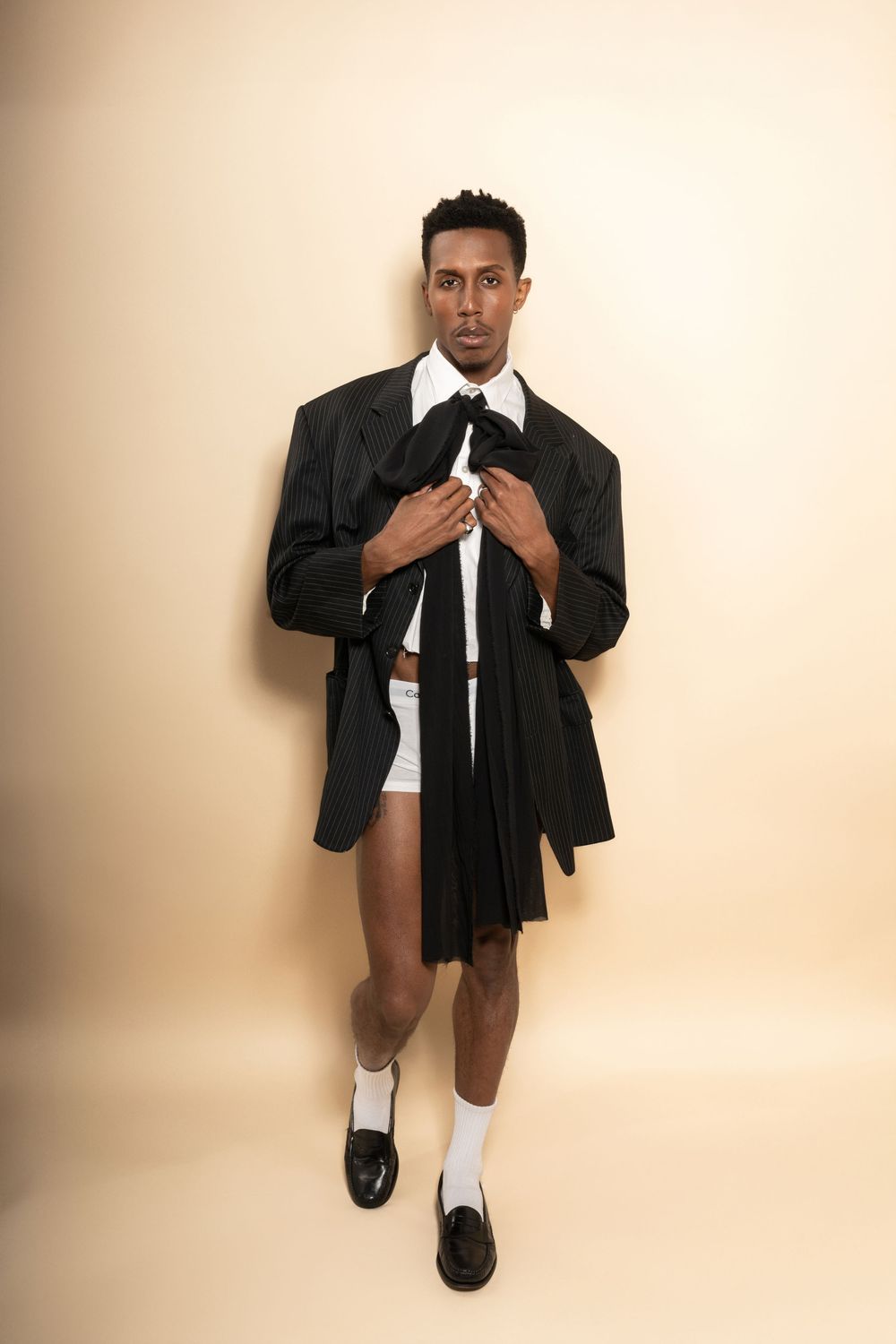 Person wearing black blazer with white shirt and tie poses in contrast with short dress code style on beige background.
