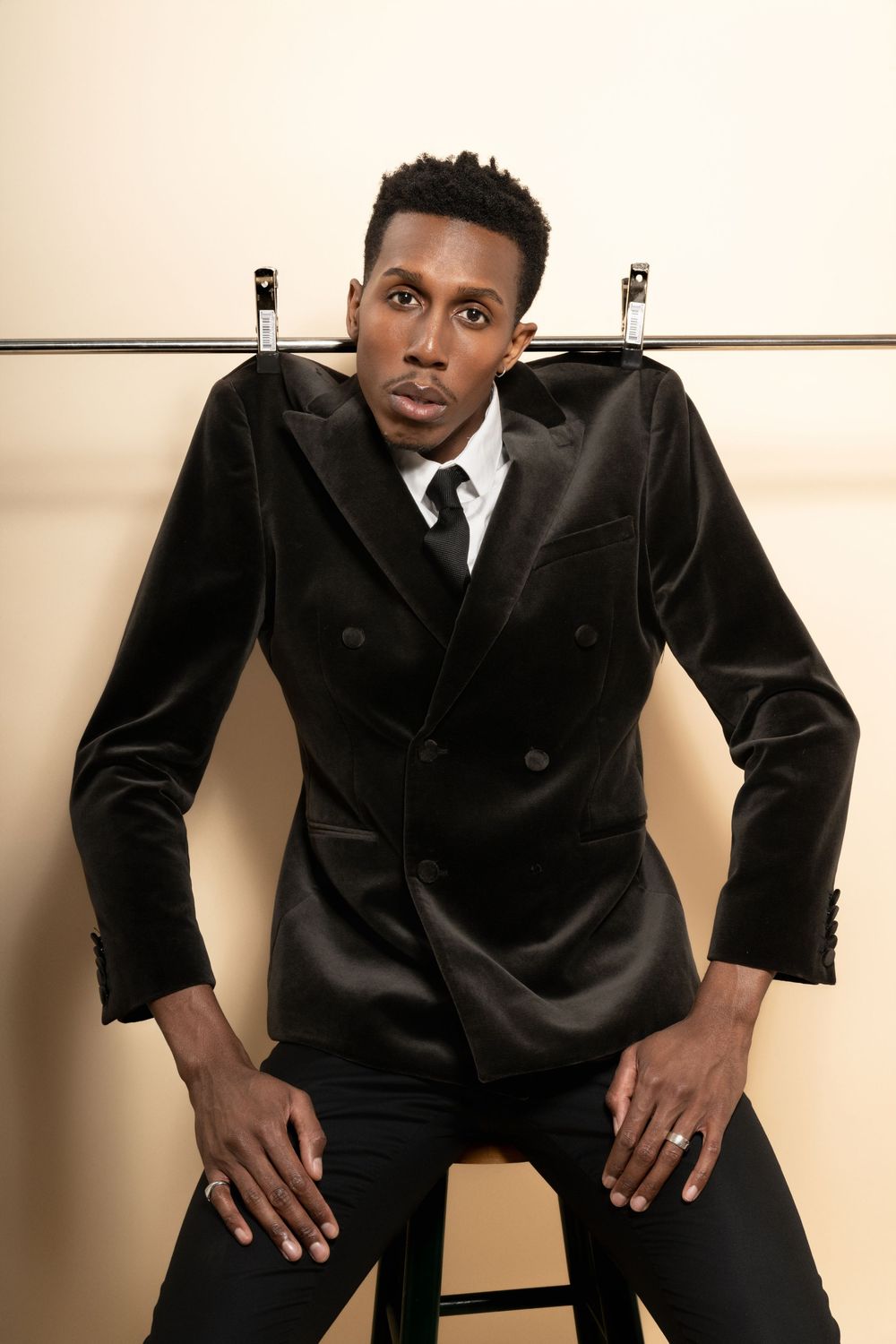 Model in luxurious black velvet double-breasted suit jacket poses on stool against neutral background.