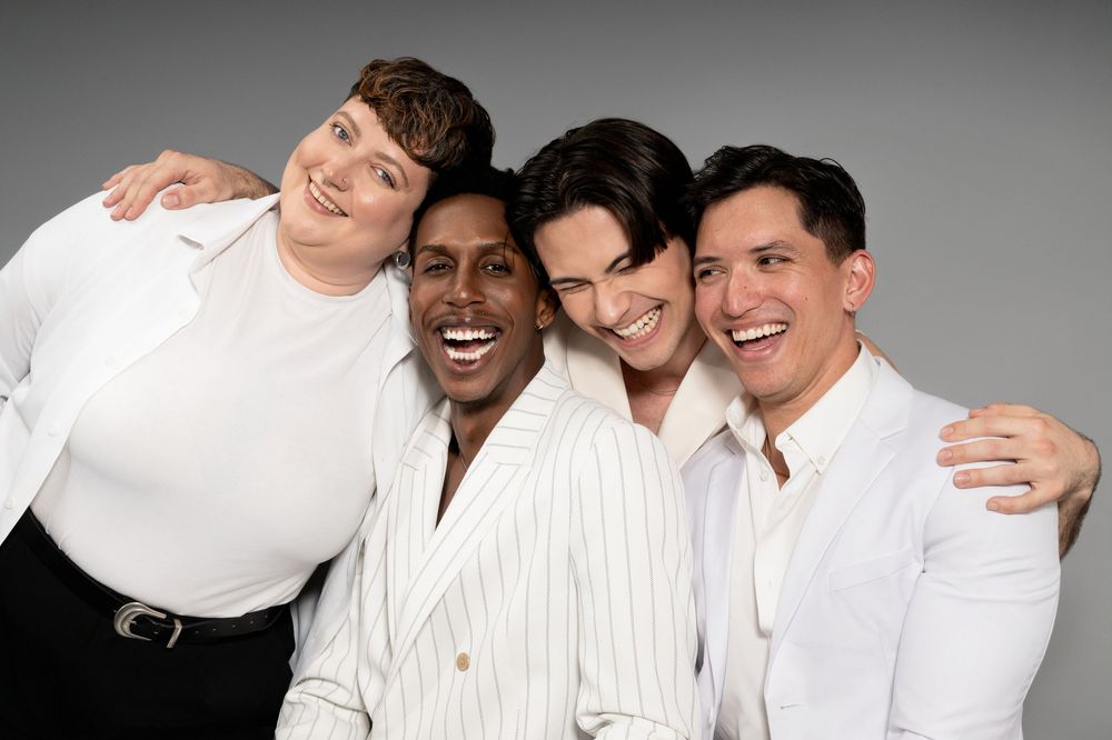 Four people wearing white clothing pose together while laughing and embracing against a gray studio background.