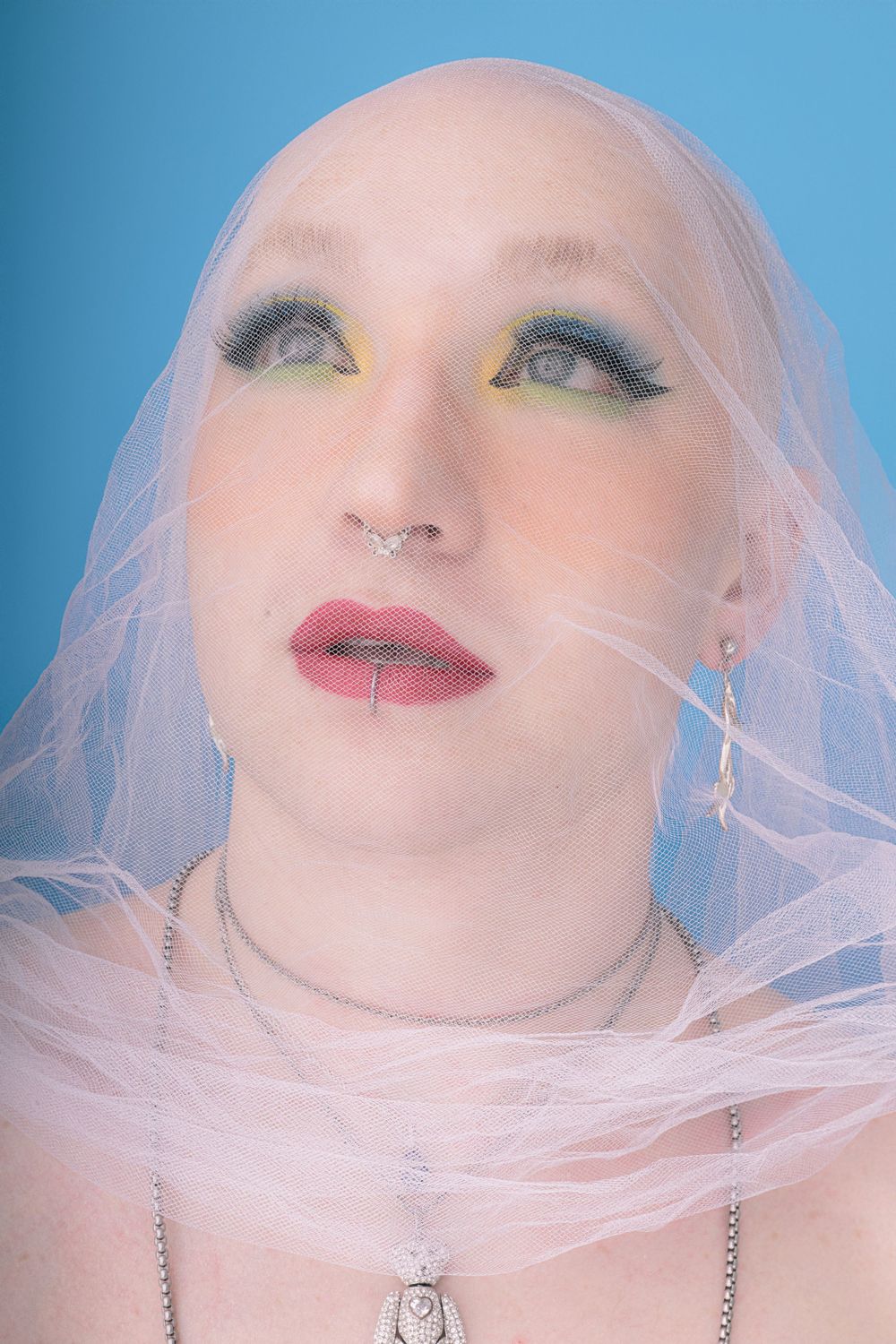 A close up portrait with dramatic makeup, pink hair and a light tulle veil against a blue background.