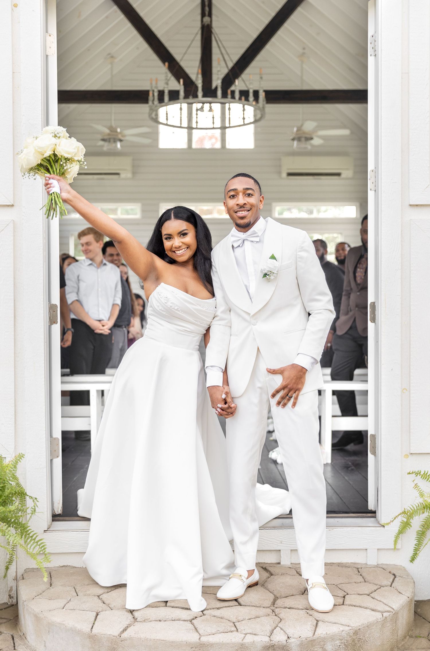 Joyful newlywed couple in white wedding attire celebrating at chapel entrance, bride raising bouquet.