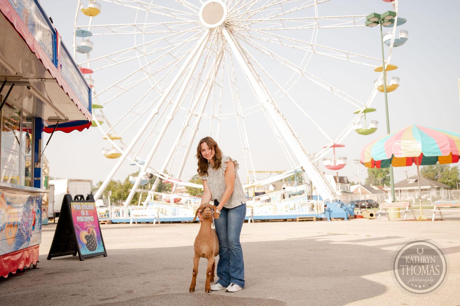 Tan goat and owner pose together near ferris wheel at carnival fairgrounds.