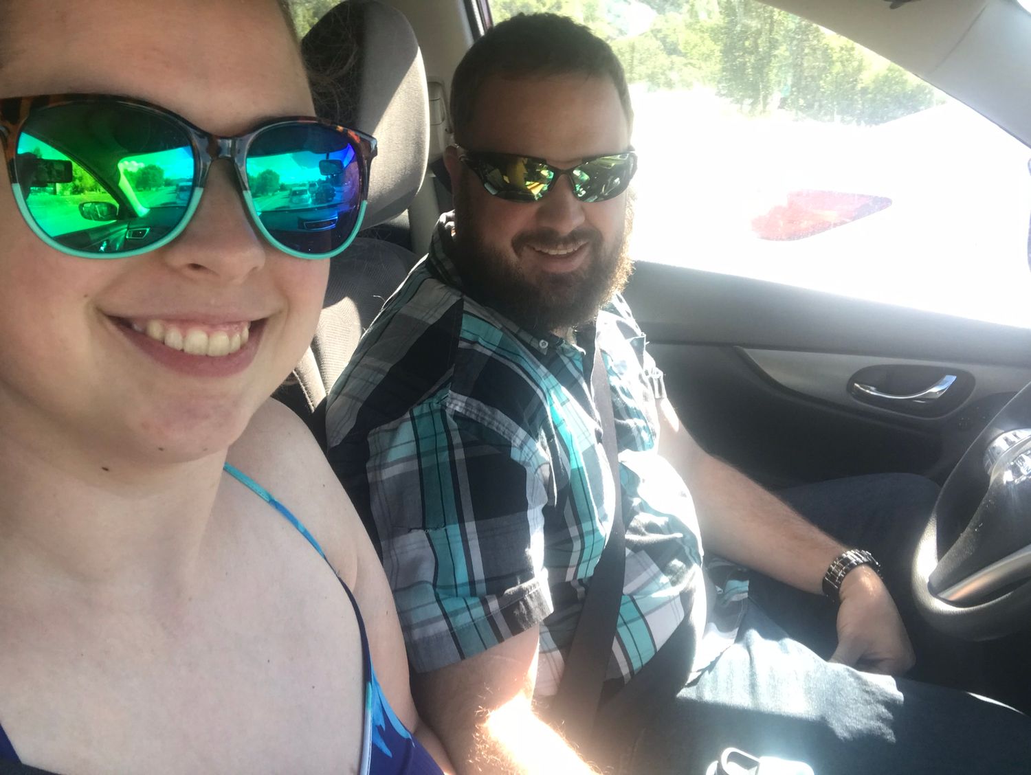 Two friends wearing reflective sunglasses taking a selfie in a car on a bright day.