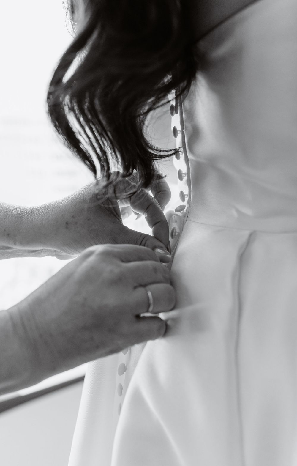 Black and white photo of hands buttoning the back of a white wedding dress on a bride with dark hair.