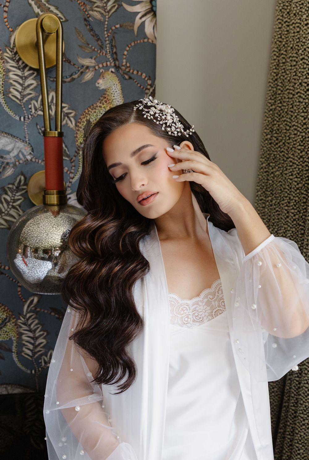 Bride in white lace robe with pearl headpiece, long wavy dark hair, posing elegantly in a decorated room.