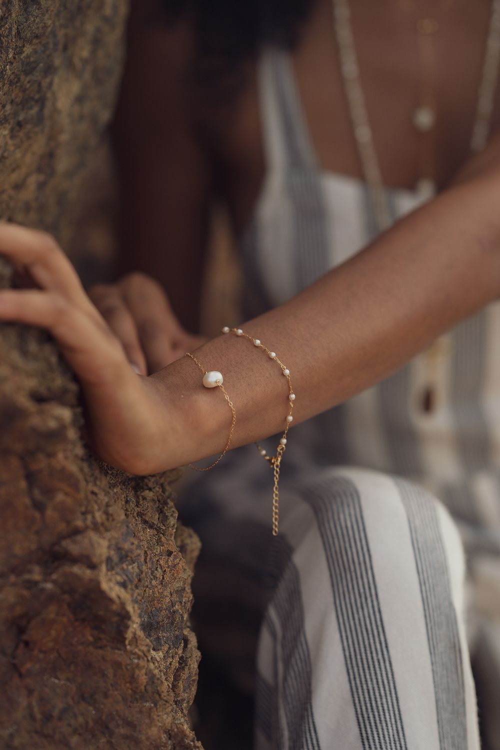 Delicate gold bracelet detail against striped fabric with rocky texture background.