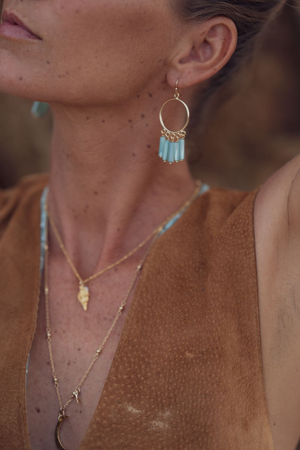 Detailed view of layered necklaces and turquoise beaded hoop earrings against a brown suede garment.