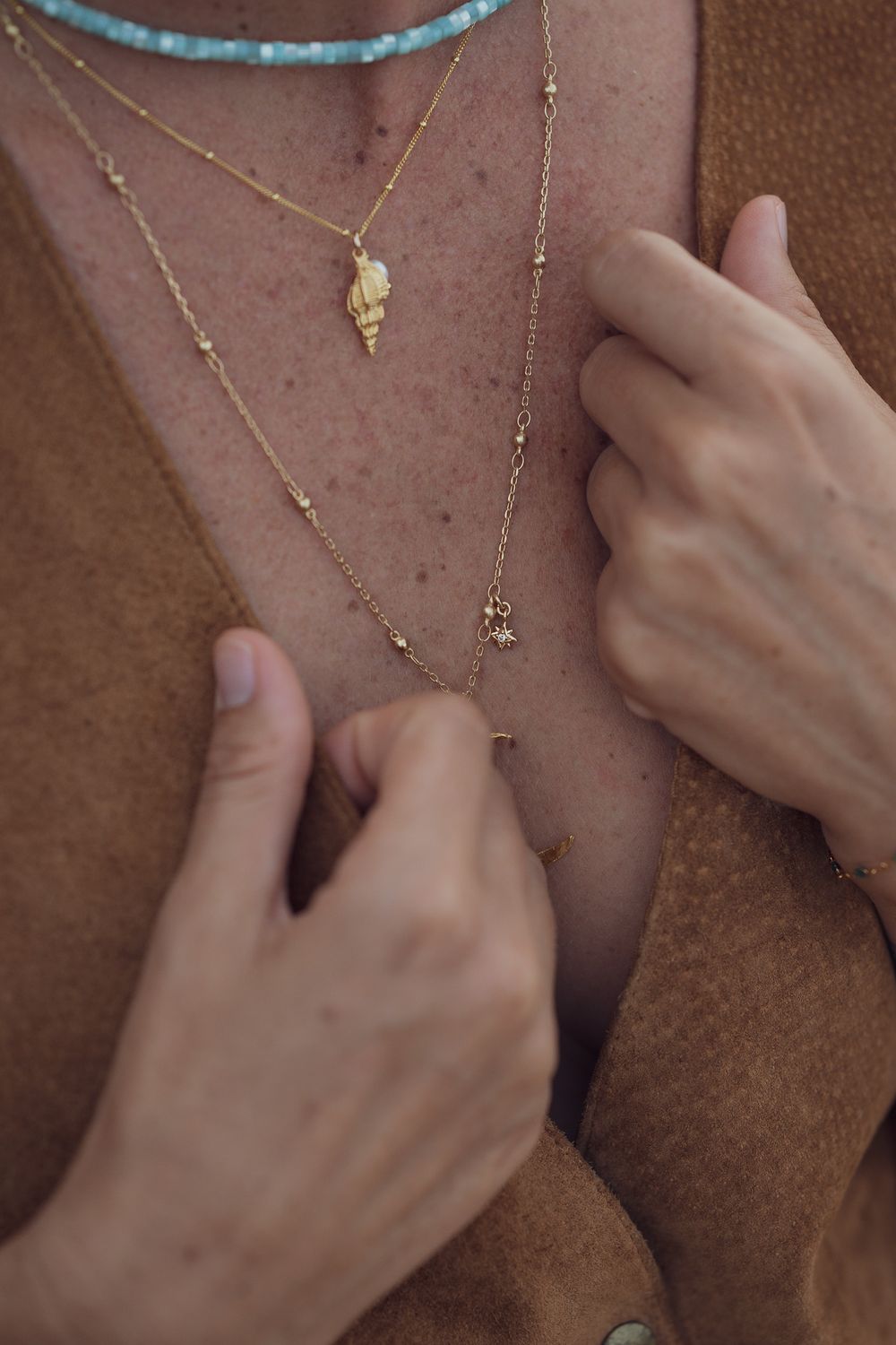 Close-up of hands adjusting delicate layered gold necklaces against a brown suede background.