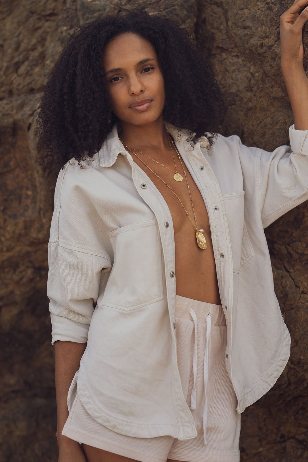 Model wearing white button down shirt and layered gold necklaces against rocky background.