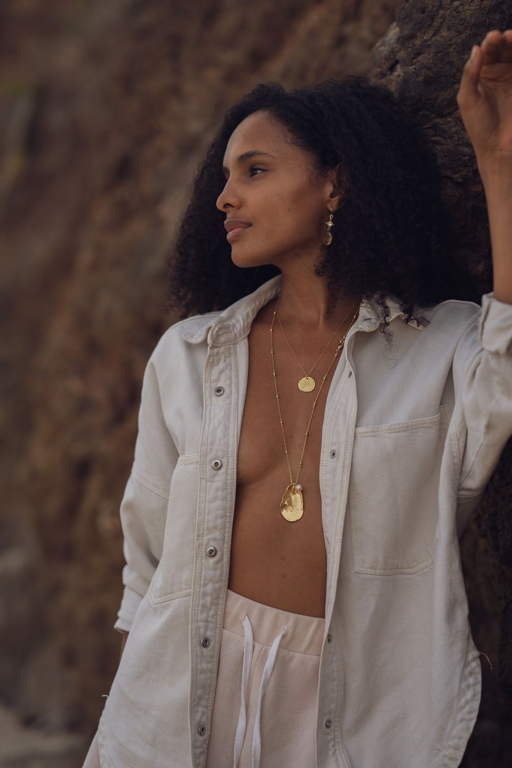 Side profile shot showing layered gold jewelry against white linen outfit and rock wall.