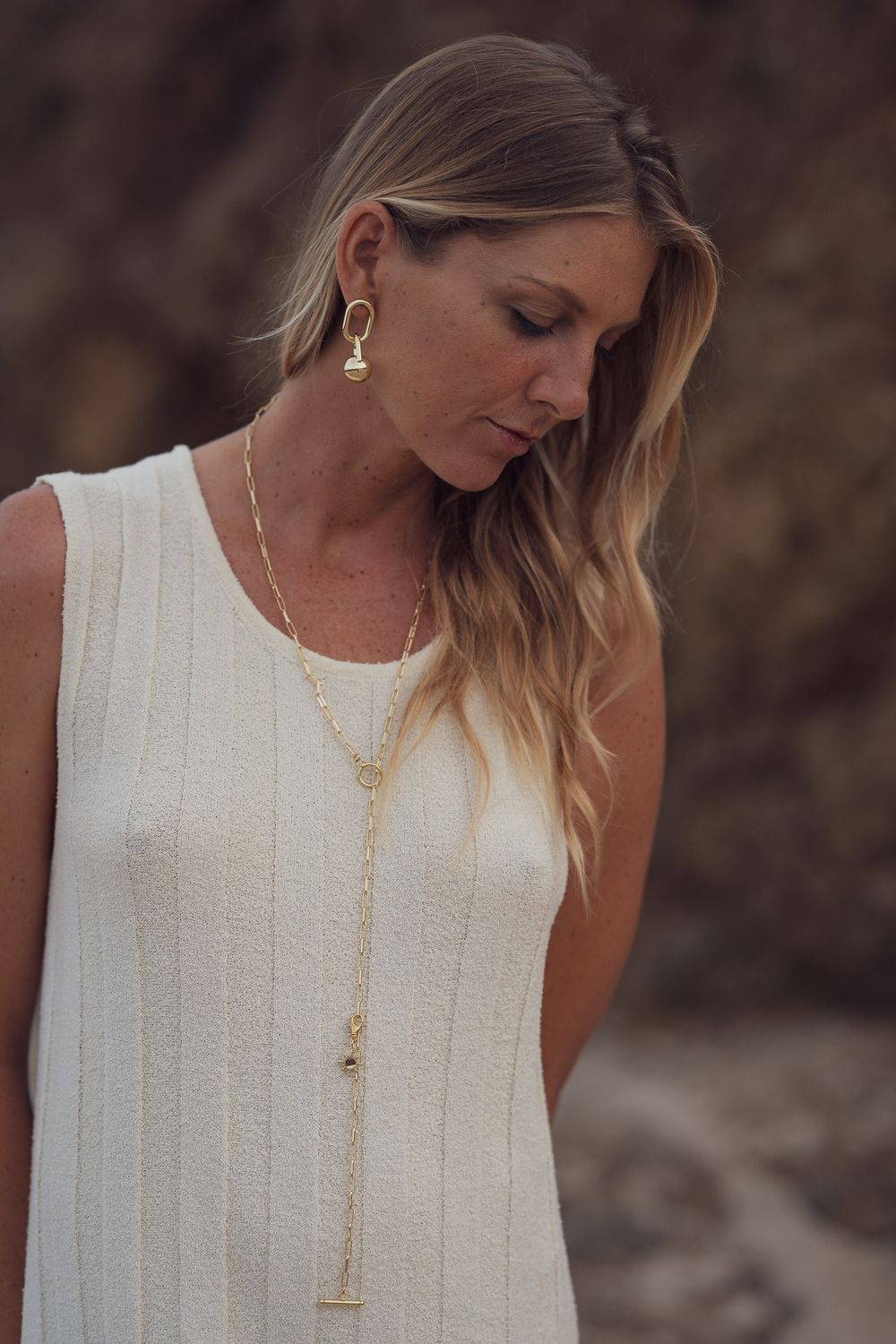 Detailed view of gold pendant necklace against white ribbed dress with natural rock backdrop.