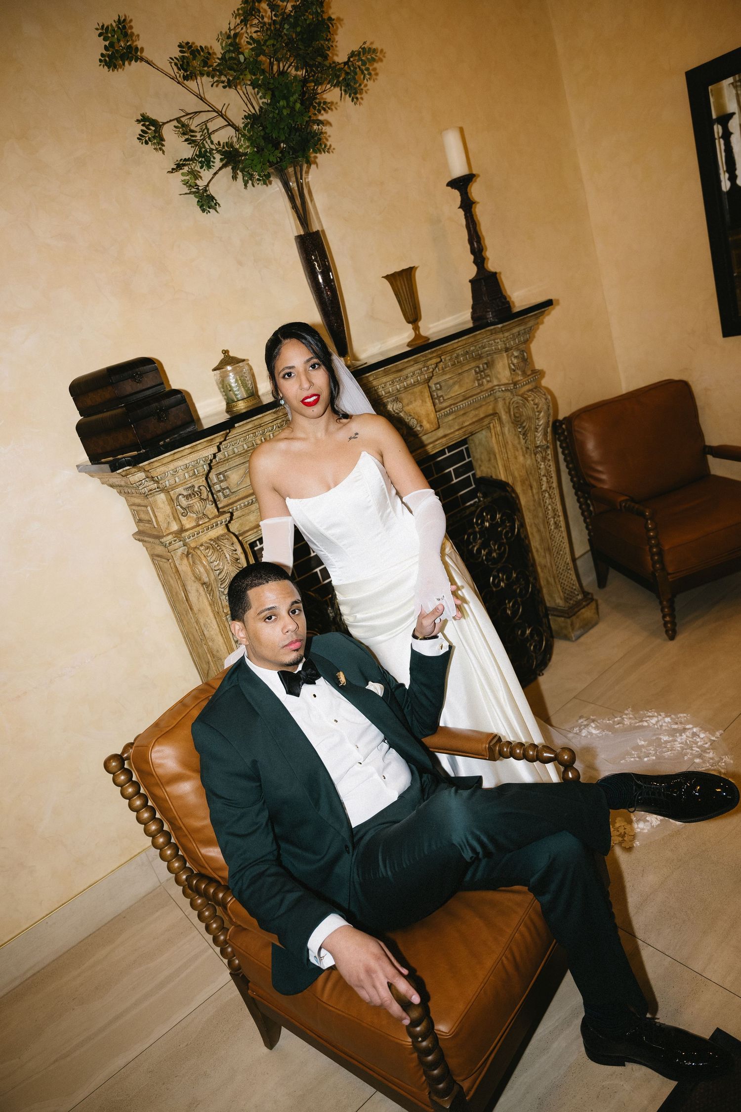 Elegant bride in white gown stands behind groom in black tuxedo seated in leather chair by ornate fireplace.