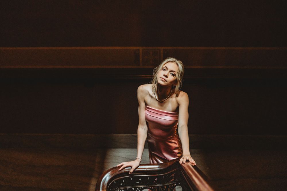 Model in pink satin dress poses elegantly in a dark interior setting.