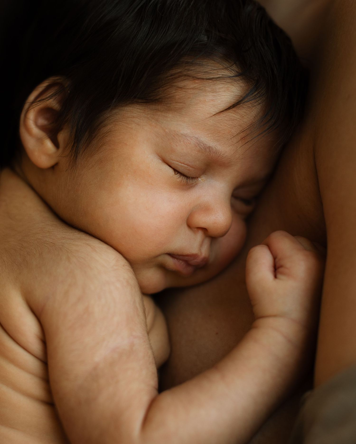 Nyfødt baby med mye svart hår som ligger hud mot hud inntil brystet på mamman sin og sover