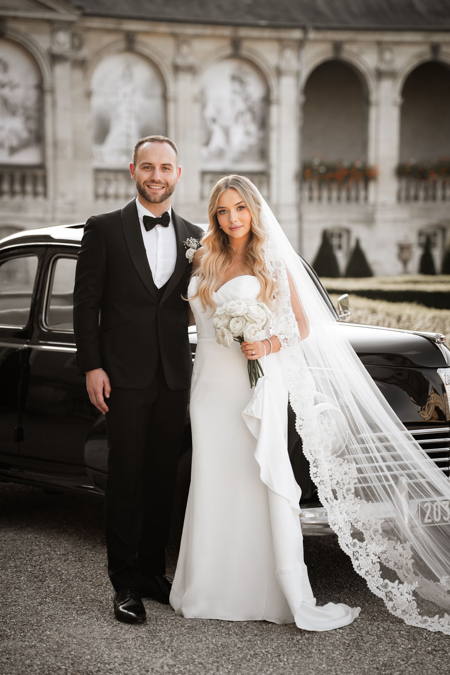 Elegant bride and groom posing in wedding attire beside a vintage black car at ornate venue.