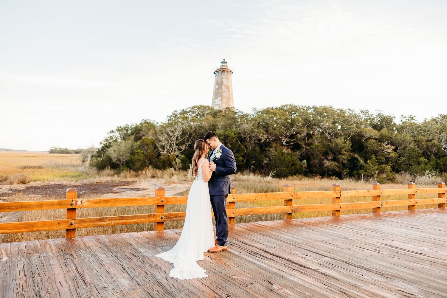 A couple shares a romantic moment on a wooden boardwalk at sunset with a lighthouse in the distance.