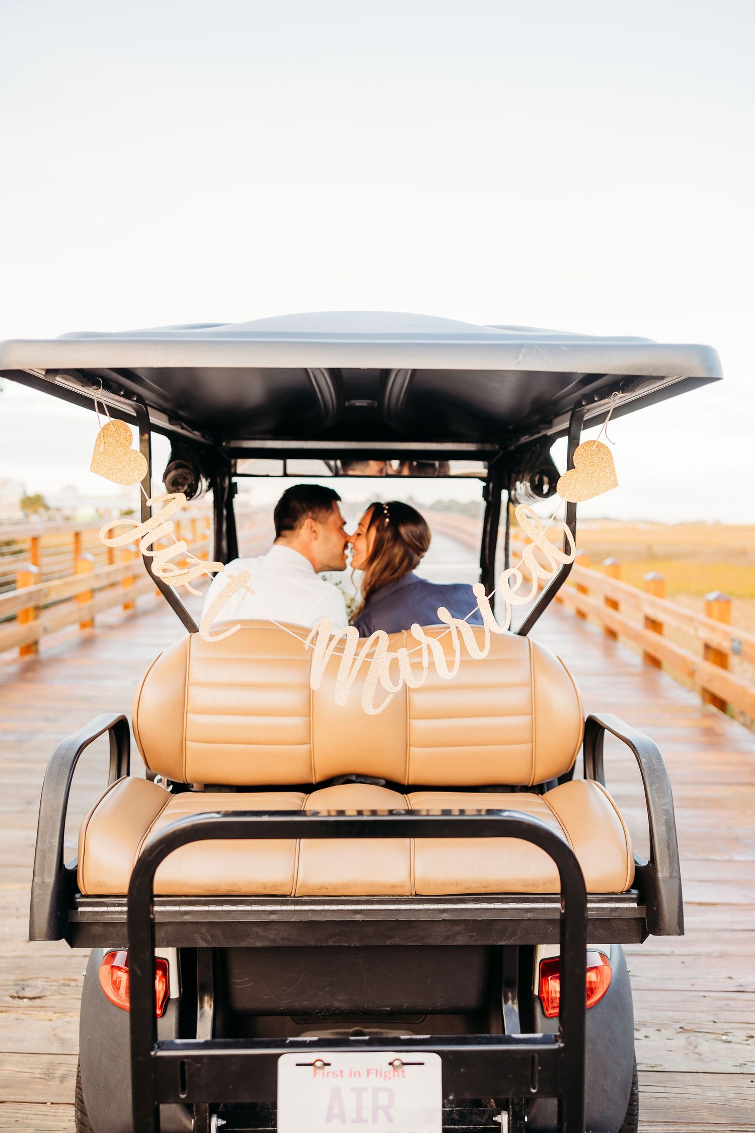 A couple sits in a golf cart on a wooden boardwalk at sunset, creating a romantic silhouette against the sky.