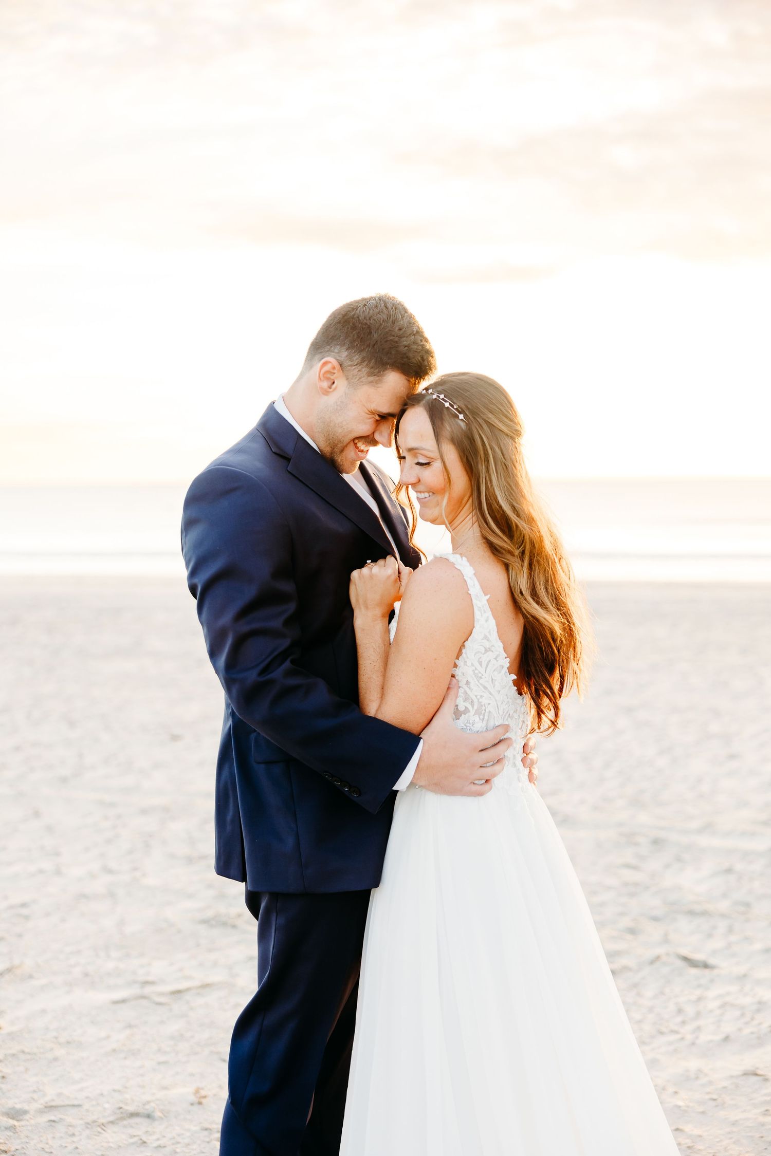 A romantic couple in wedding attire share an intimate moment on a sandy beach at sunset.