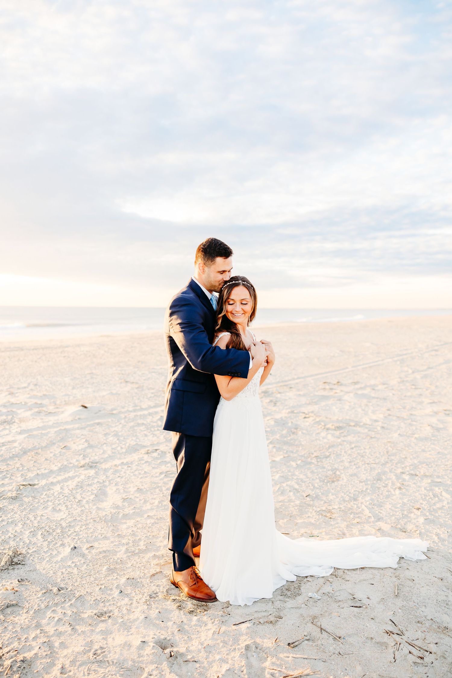 A couple in wedding attire embraces on a scenic beach during sunset with soft golden light illuminating the sand.
