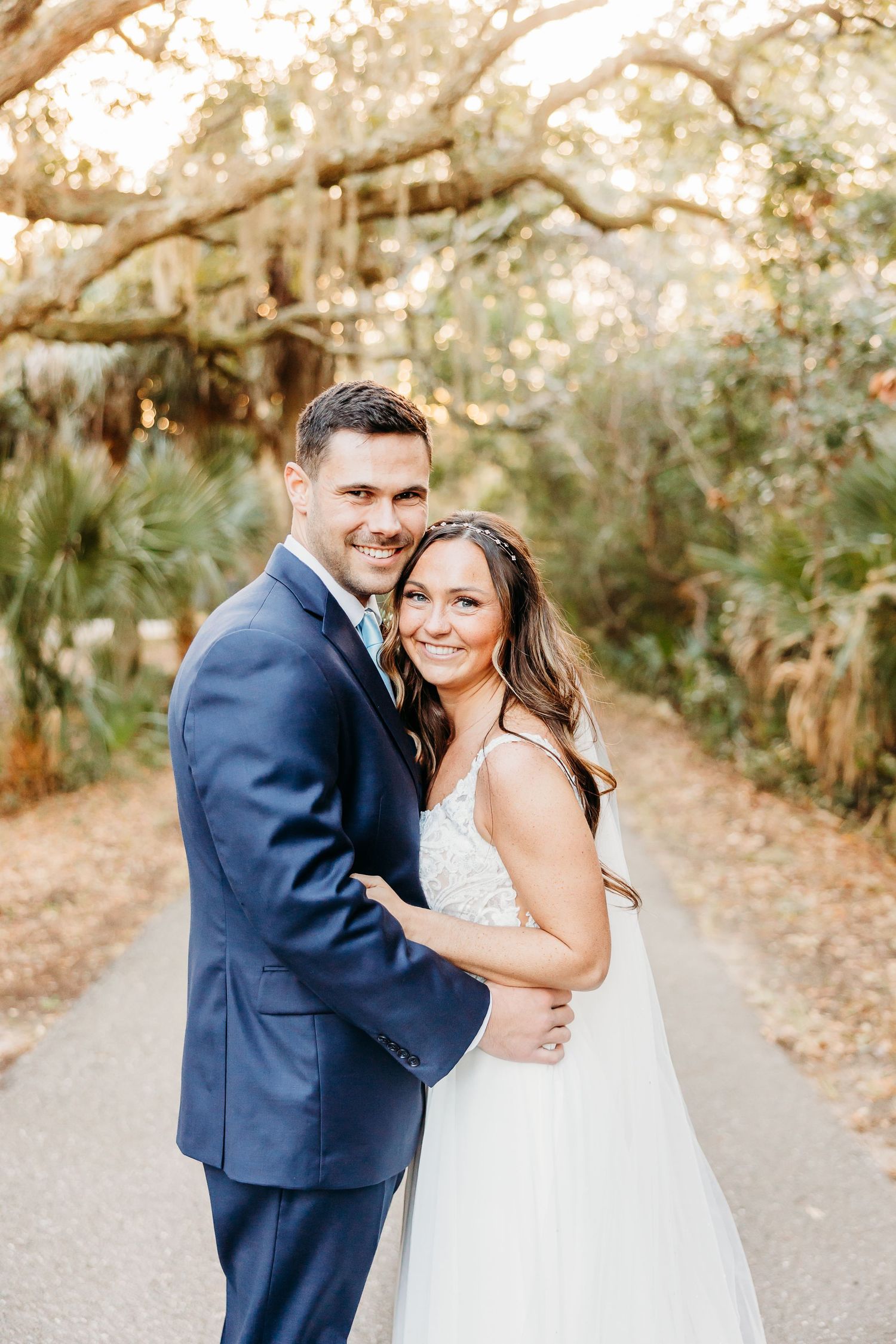 A bride and groom share a joyful moment together on a tree-lined path during their wedding photoshoot.
