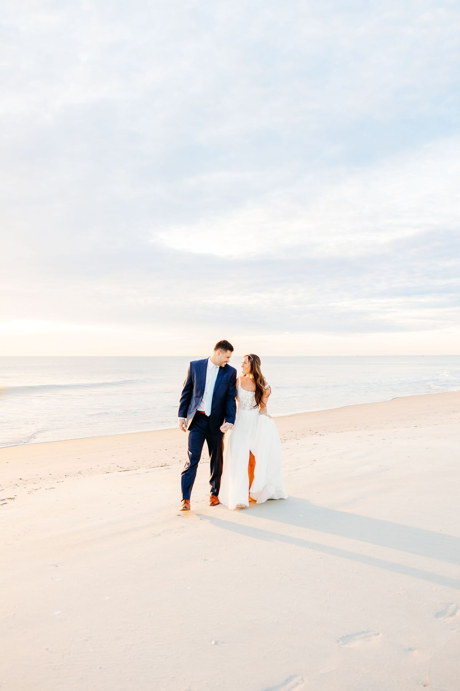 A couple walks hand in hand along a serene beach at sunset with soft pastel colors in the sky.