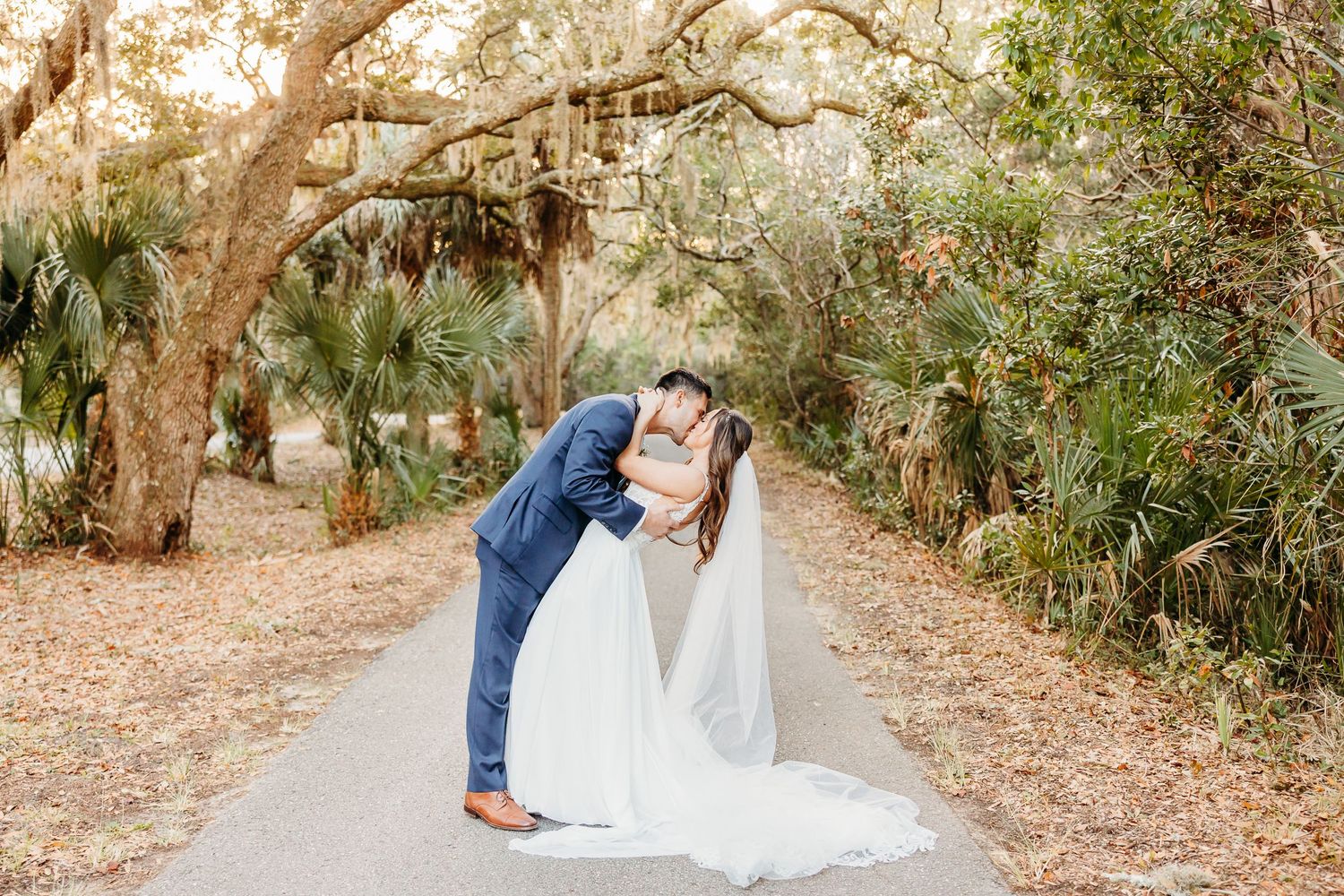 A romantic couple embraces on a sunlit path surrounded by trees and autumn foliage.