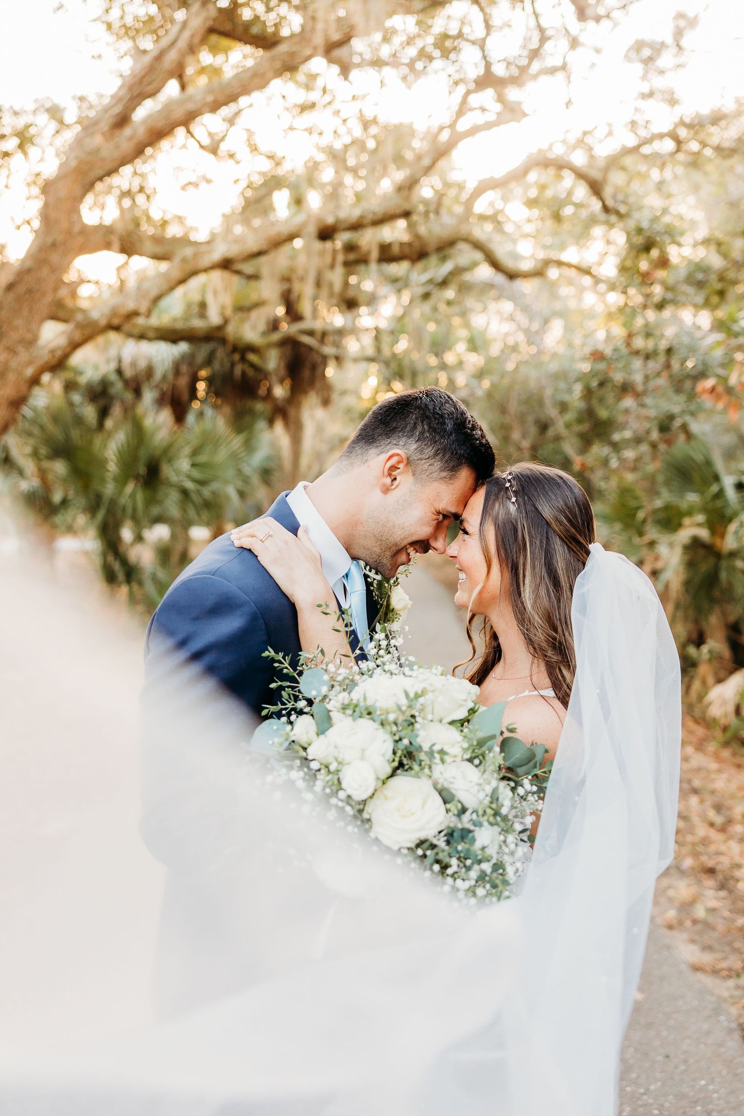 Romantic wedding couple shares intimate moment under Spanish moss draped trees with white floral bouquet and flowing veil.
