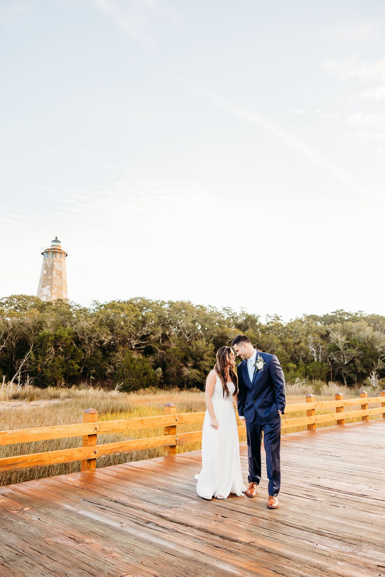 Couple walking along wooden boardwalk with lighthouse and coastal vegetation in background during golden hour.