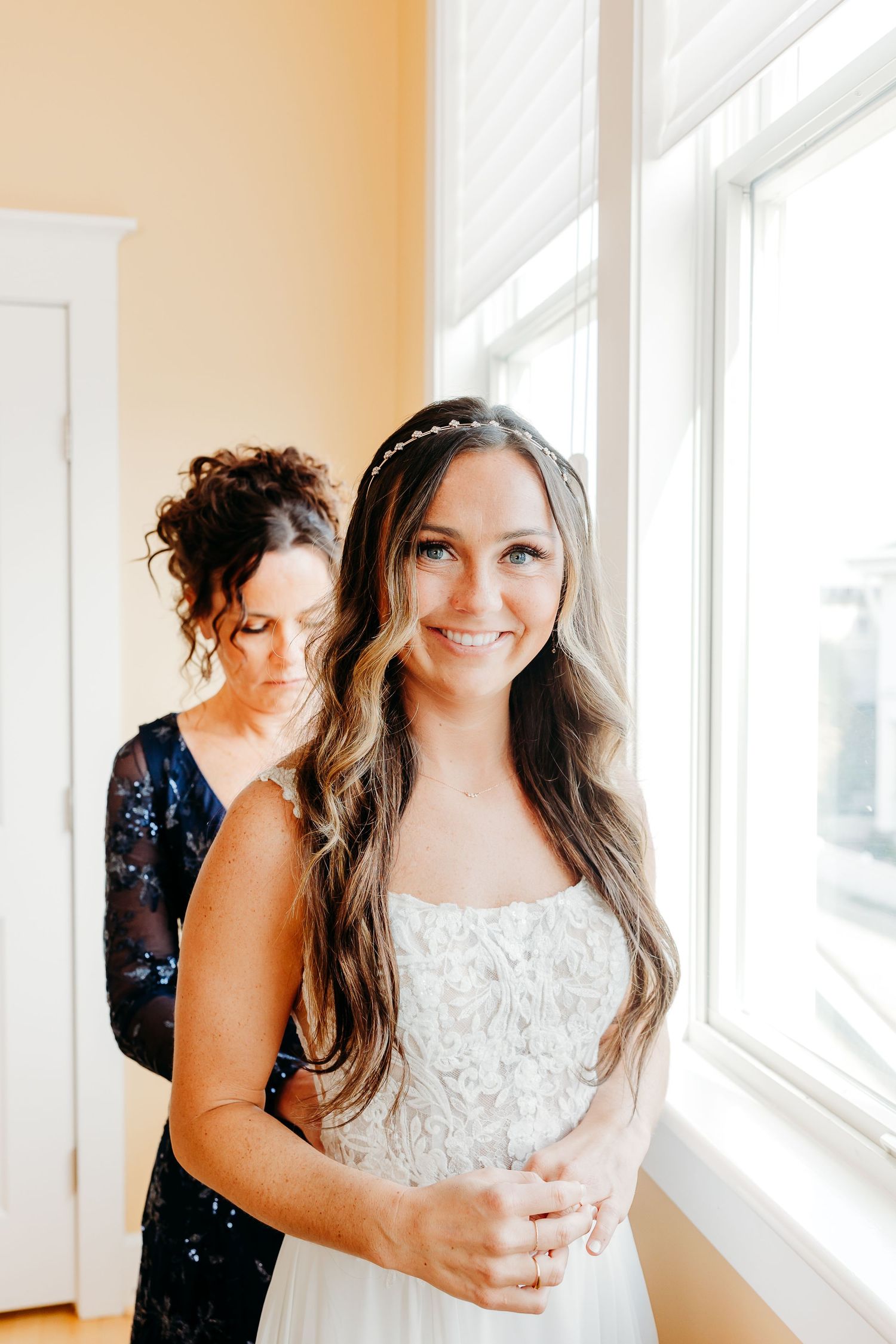 A bride in a white lace dress smiles brightly near sunlit windows during wedding preparations.