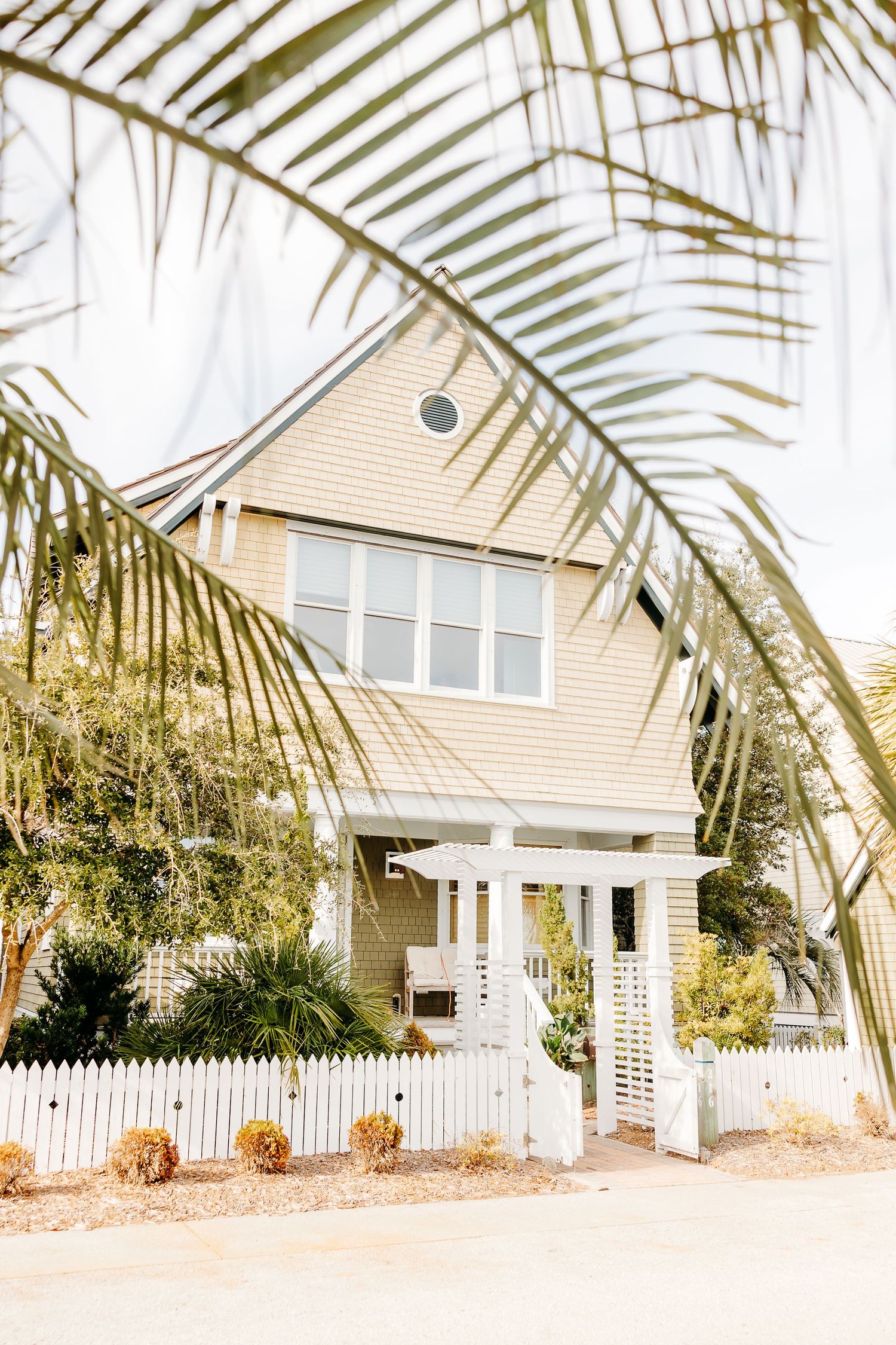 A beige coastal cottage with white picket fence framed by palm fronds and decorated with pumpkins by the entrance.