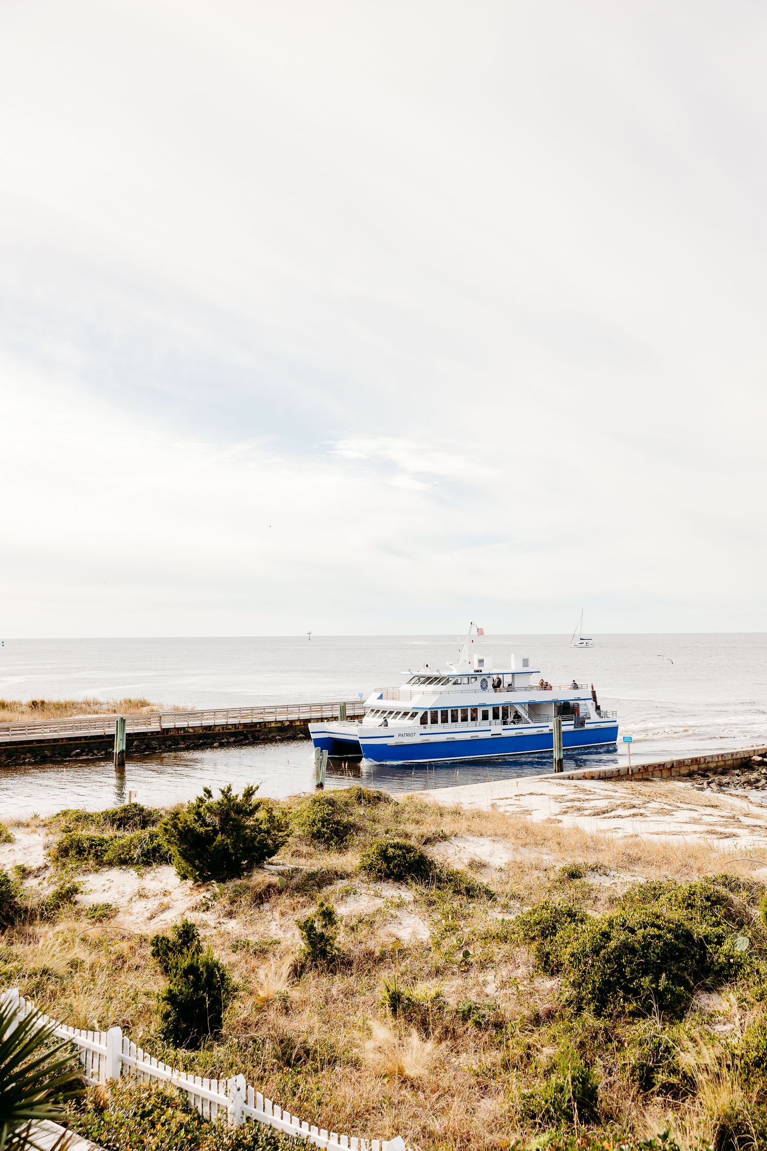 A blue passenger ferry boat docked at a coastal pier with sandy dunes and beach grass in the foreground.