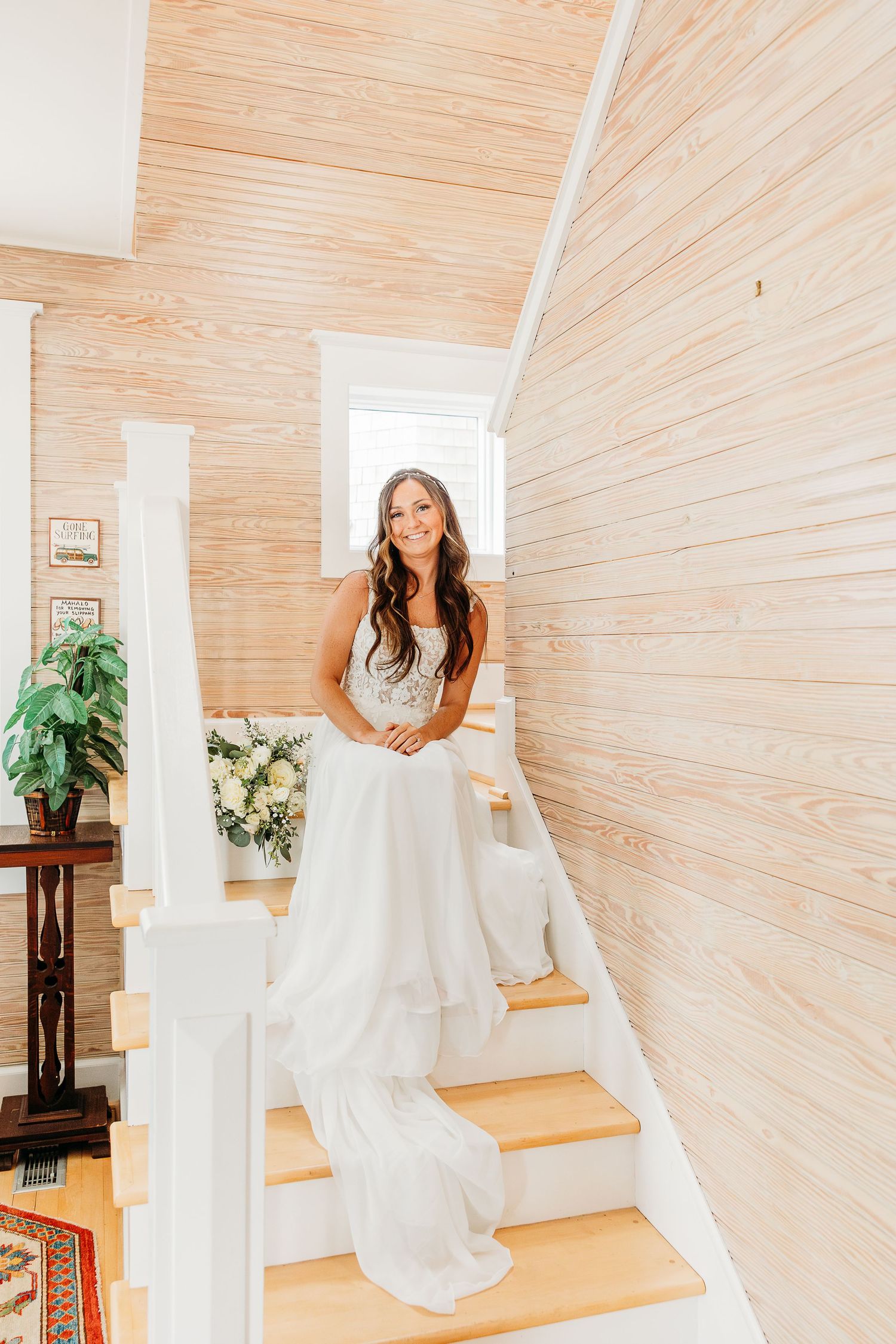 A bride in a flowing white wedding gown poses gracefully on wooden stairs with light wood paneled walls.