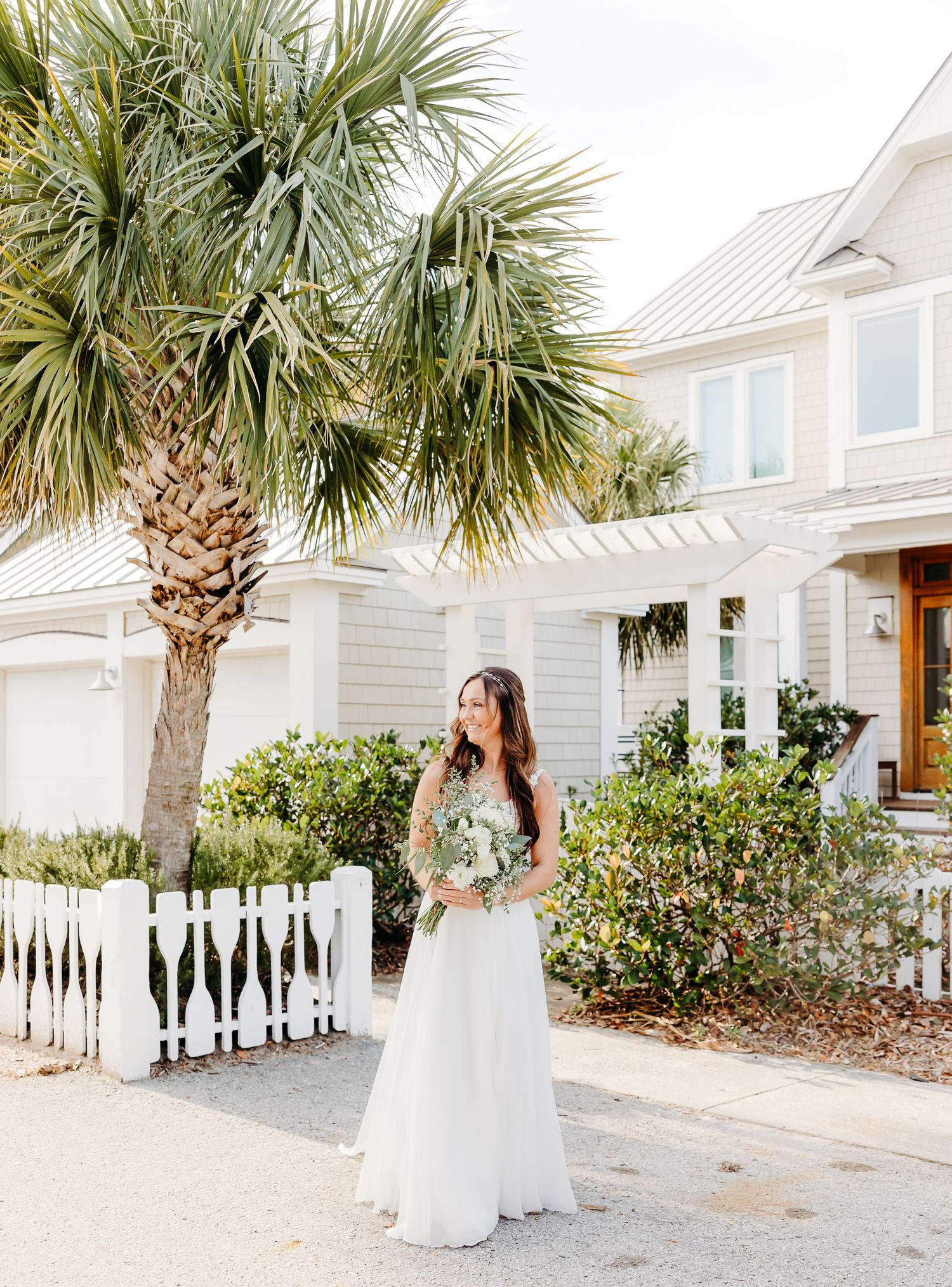 A bride in a white wedding dress stands under palm trees outside a coastal beach house with white siding.