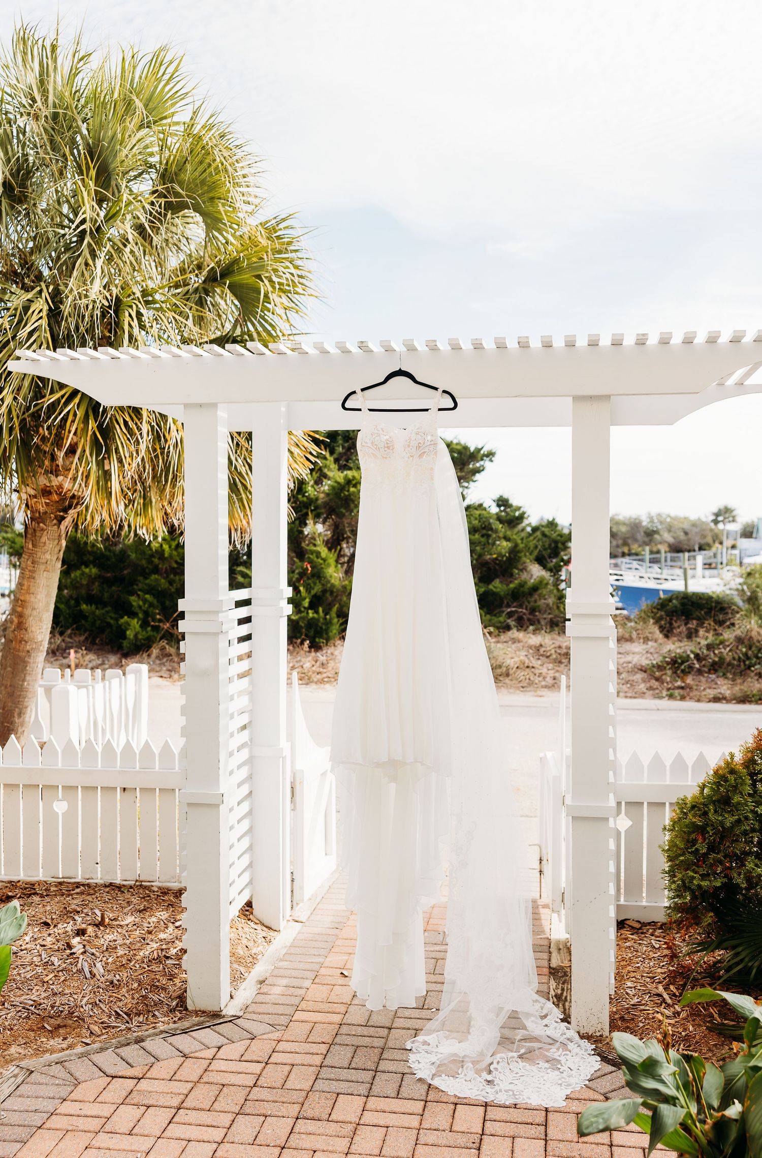 White wedding dress hanging from a pergola on a beach boardwalk with palm trees and coastal vegetation.