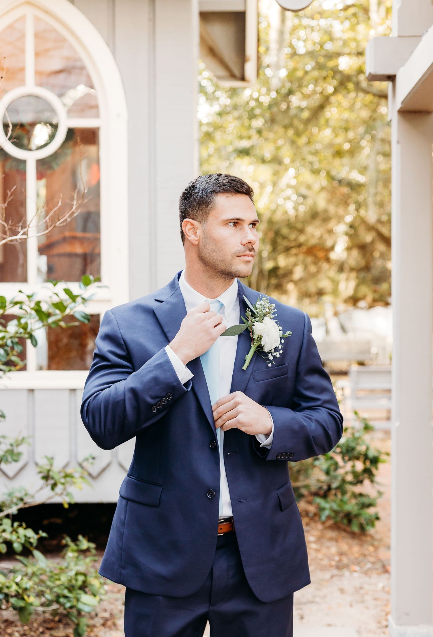 A person in a navy suit adjusts their tie while standing outside near a white porch with autumn foliage in the background.