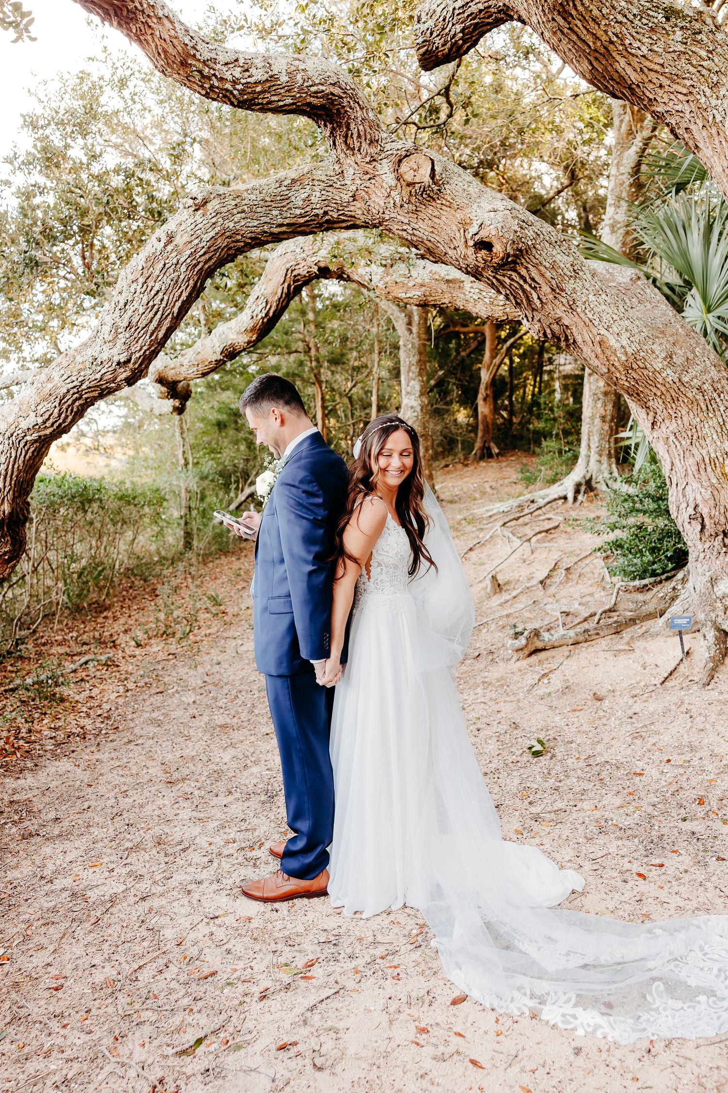A romantic wedding couple shares intimate moments under twisted oak trees on a natural dirt path in golden sunlight.