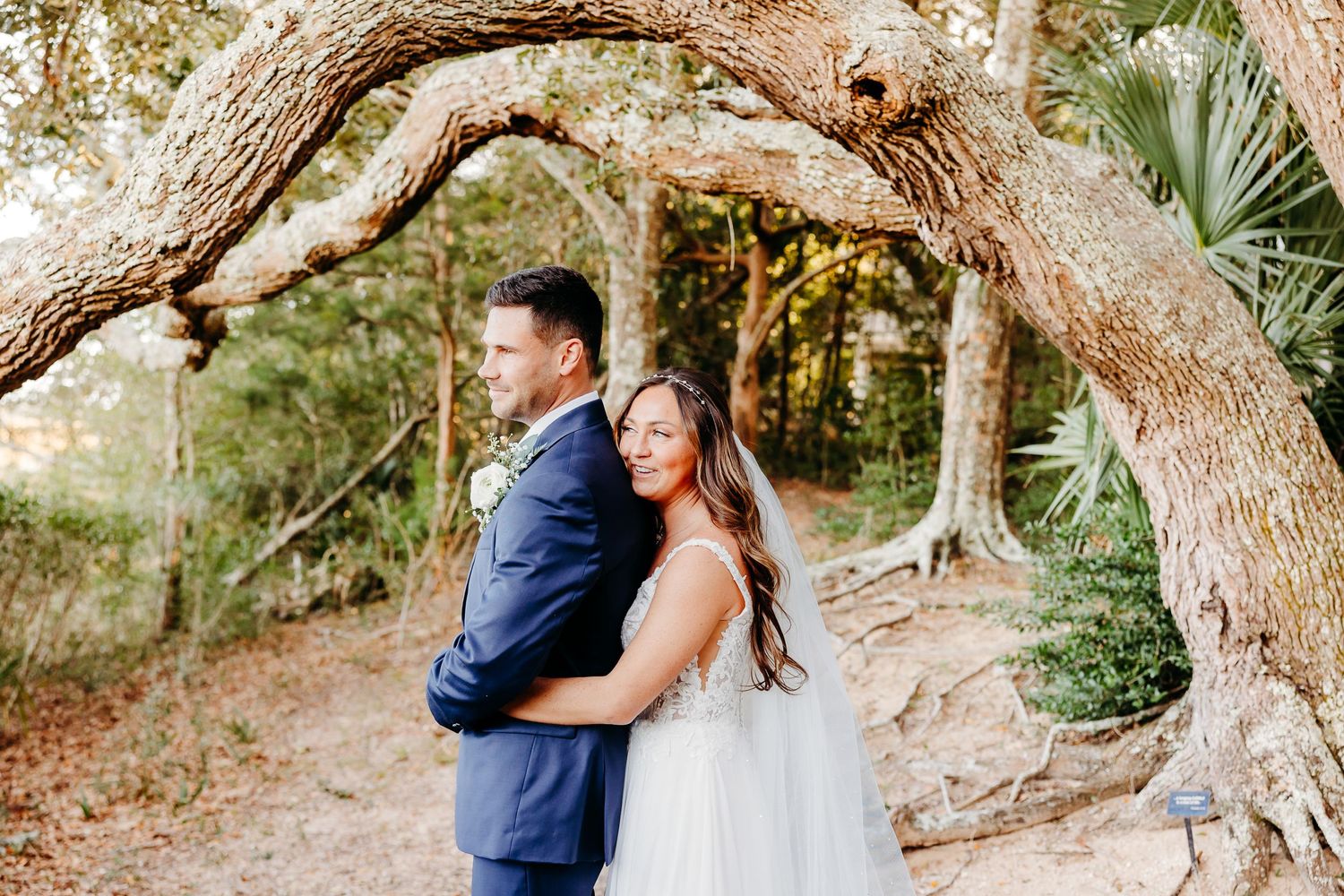 A couple in wedding attire shares an intimate moment under large tree branches in a natural outdoor setting.