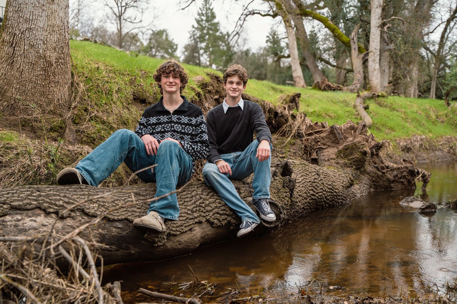 Two people sitting casually on fallen log over stream in wooded outdoor setting.