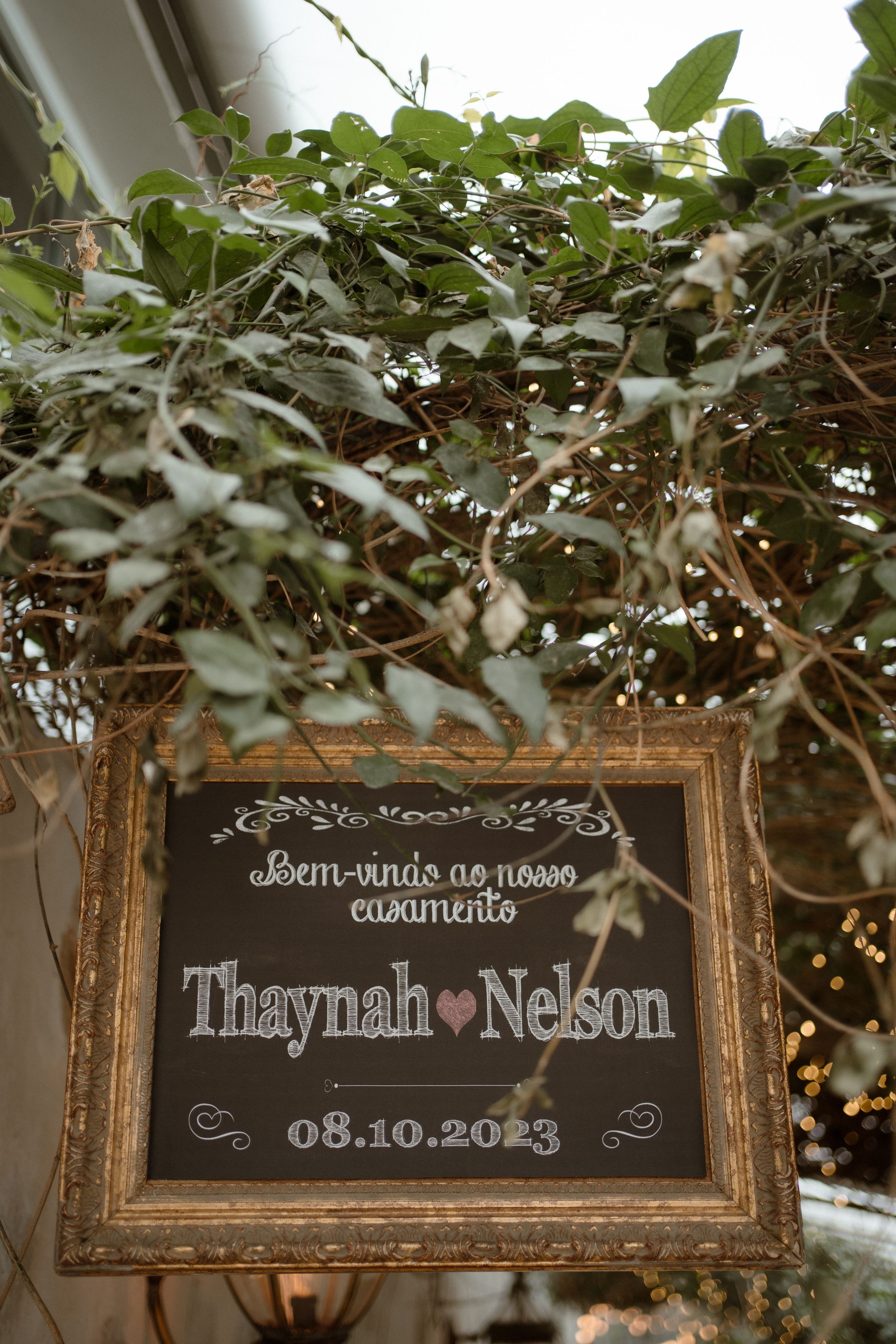 Rustic wedding welcome sign with names and date framed by greenery and twinkle lights.