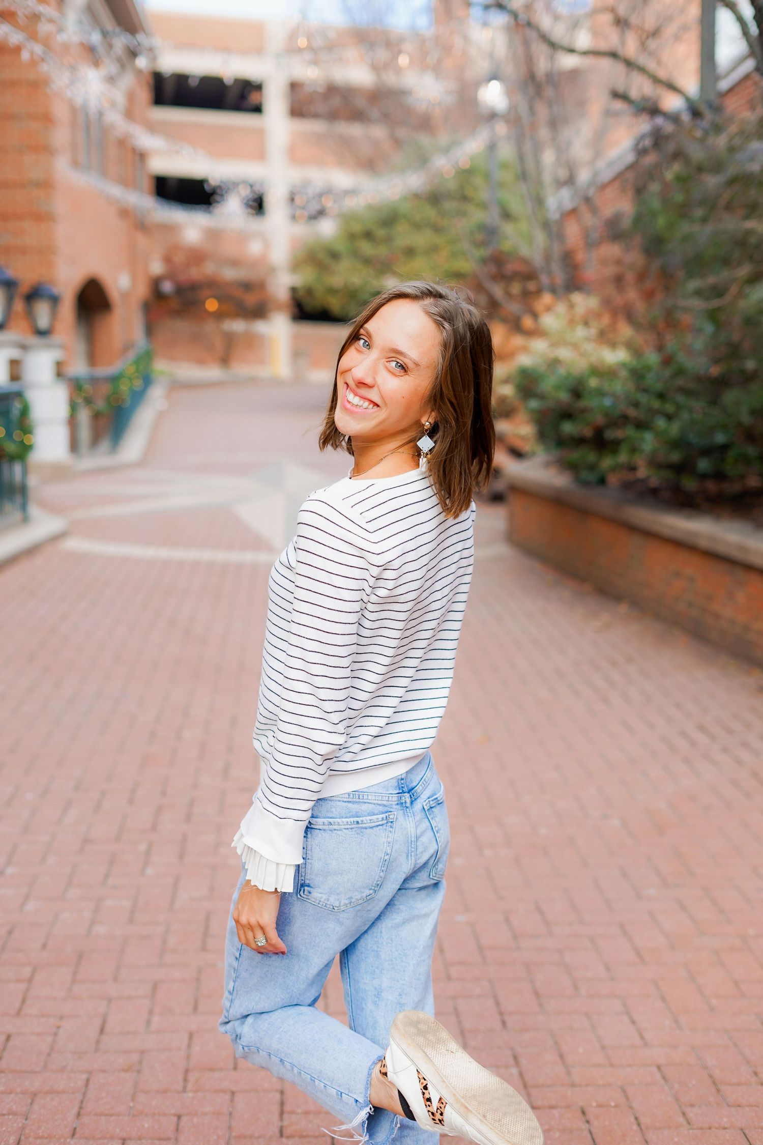 Woman in casual stripes and denim smiling on brick-paved walkway.