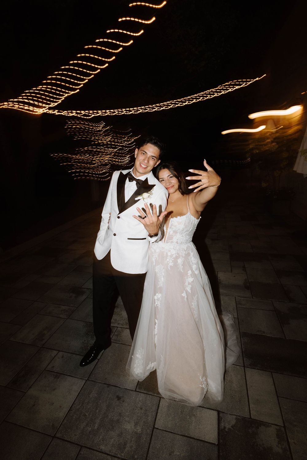 A couple dances together under romantic string lights during their wedding reception on a stone patio at night.