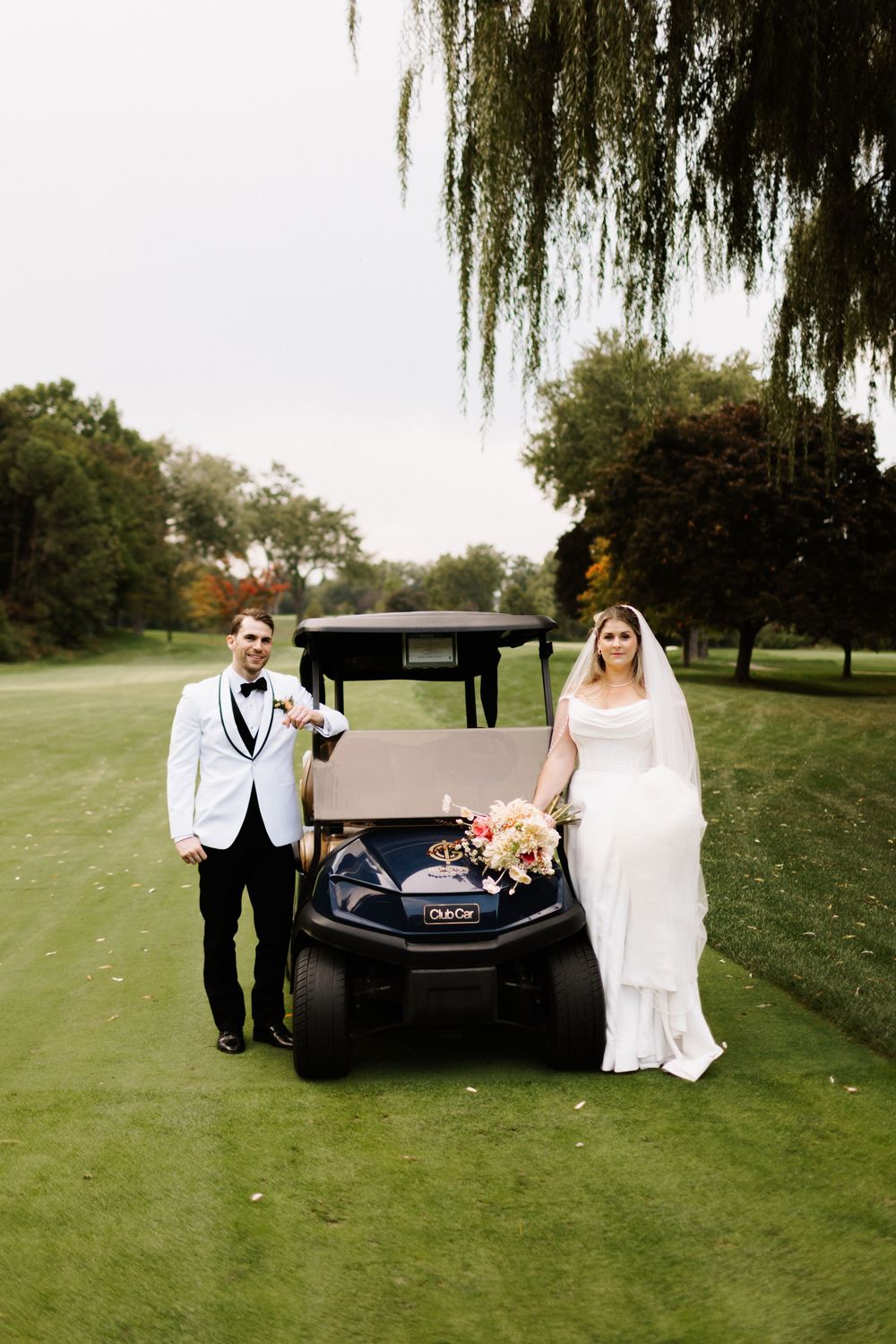 A wedding couple relaxes on a black golf cart under weeping willow trees on a grassy lawn.
